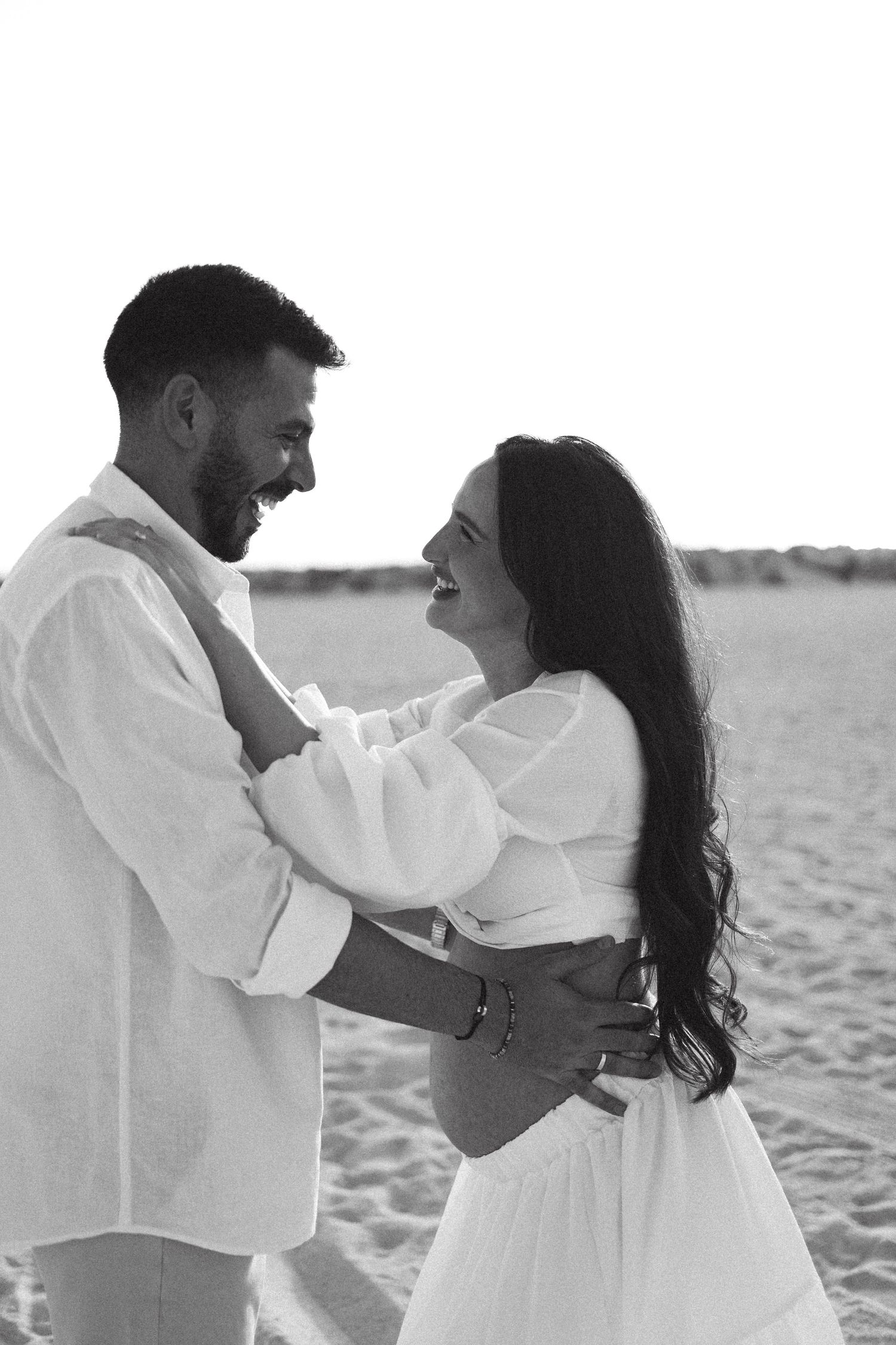 A couple in white attire dance together on a beach during sunset.