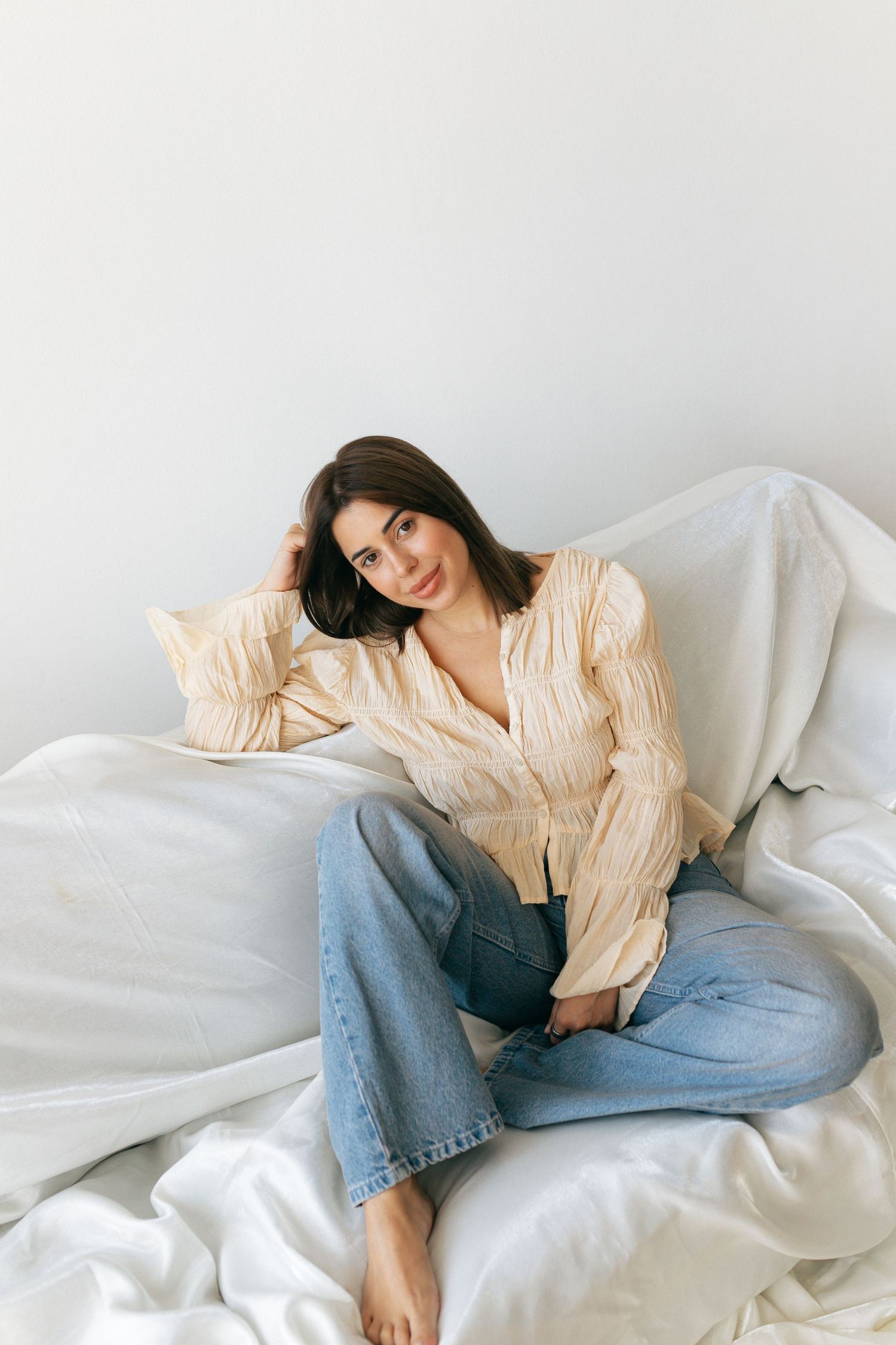Young woman in beige striped blouse and blue jeans sitting relaxed on white bedding, smiling softly at camera.