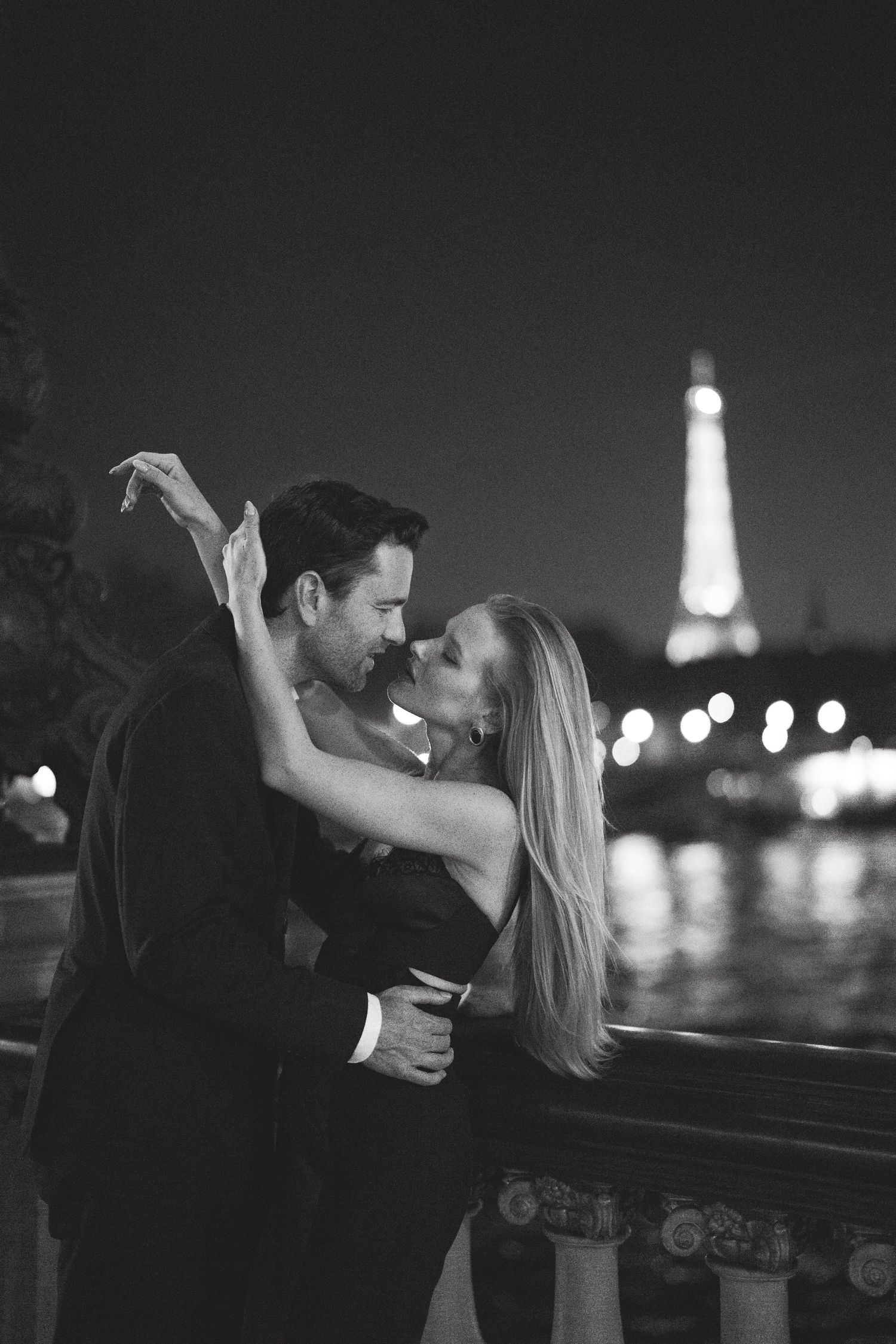 Black and white photo of a romantic couple embracing near the Eiffel Tower at night in Paris.