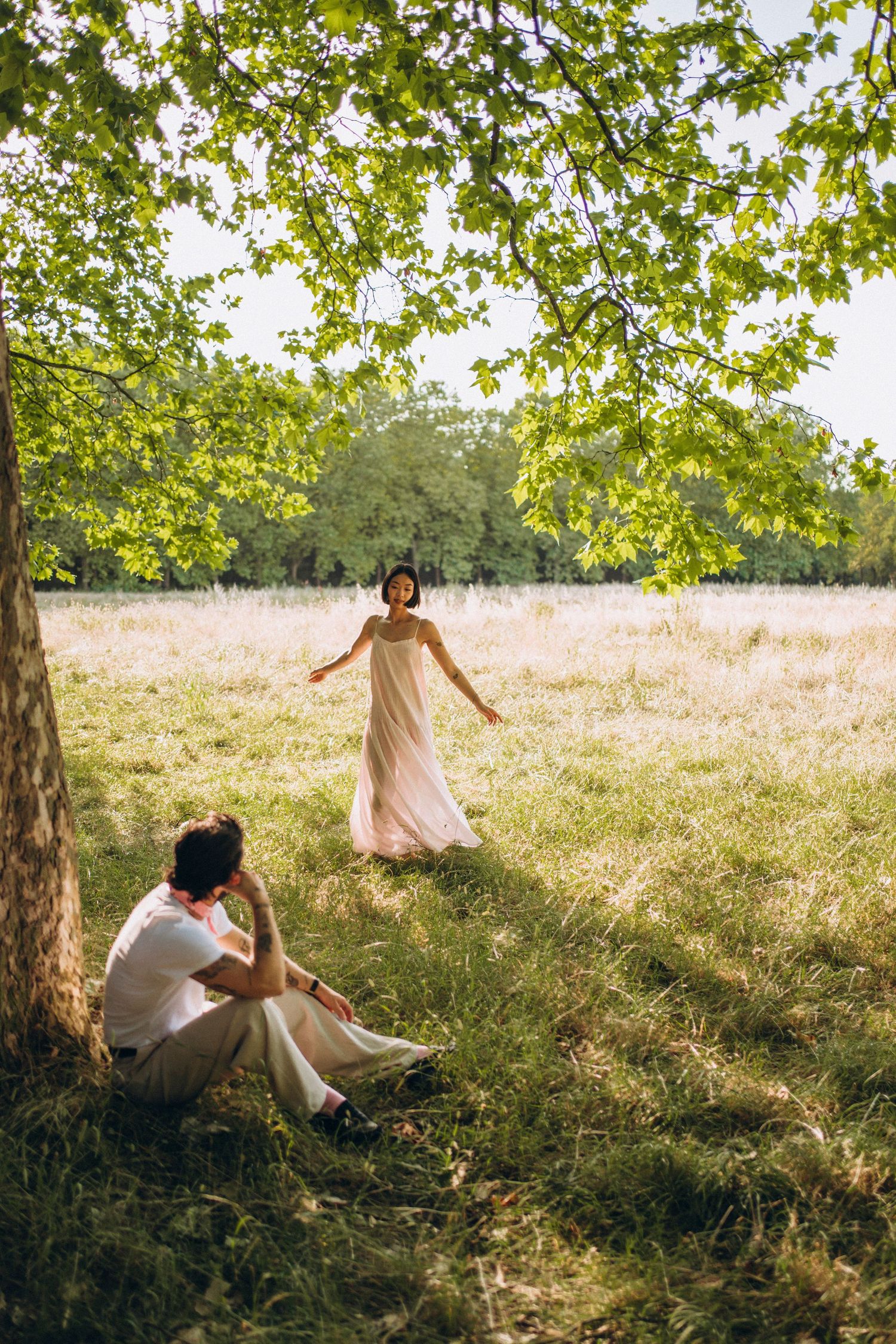 A couple in a sunlit meadow, woman in flowing pink dress walking toward man resting against a tree.