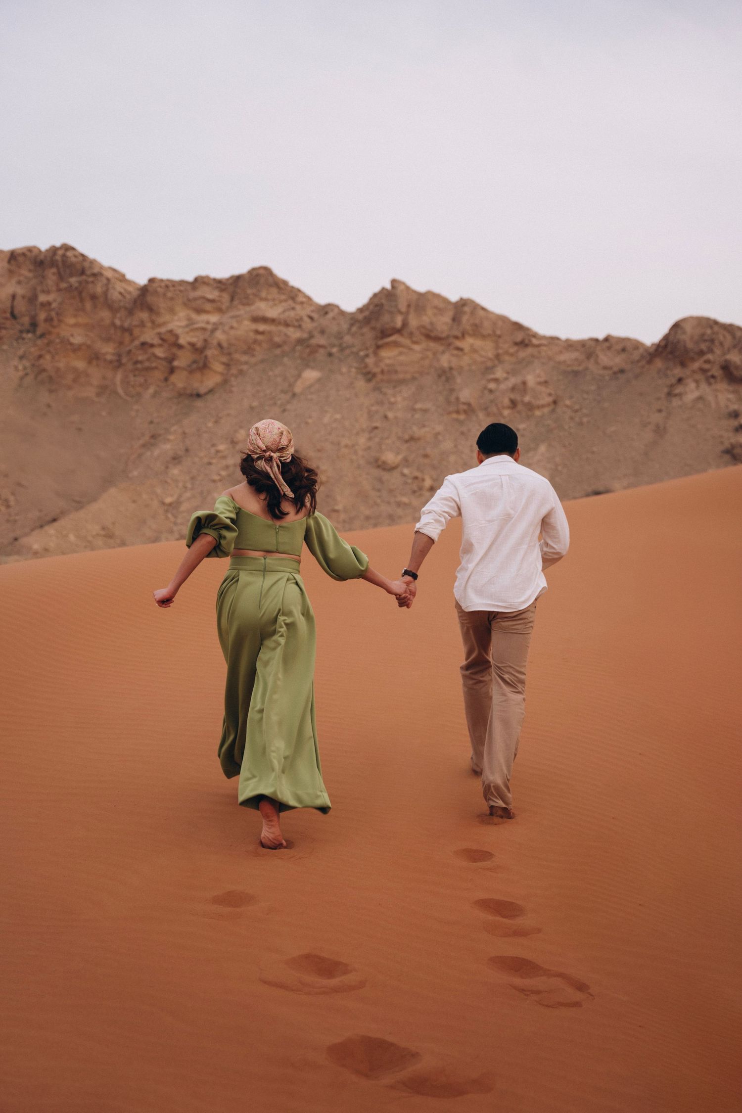 Two people holding hands while walking through desert landscape with mountain backdrop.