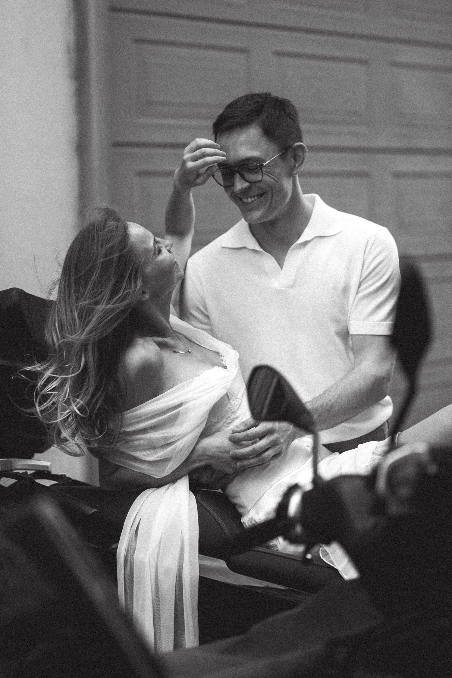 Black and white photo of a smiling couple sharing a joyful moment, man in white polo adjusting sunglasses, woman in flowing dress.