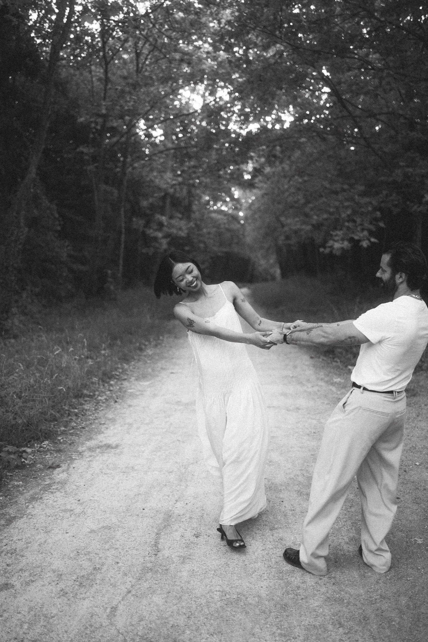 Black and white photo of a laughing couple holding hands and spinning on a tree-lined dirt path in a forest.