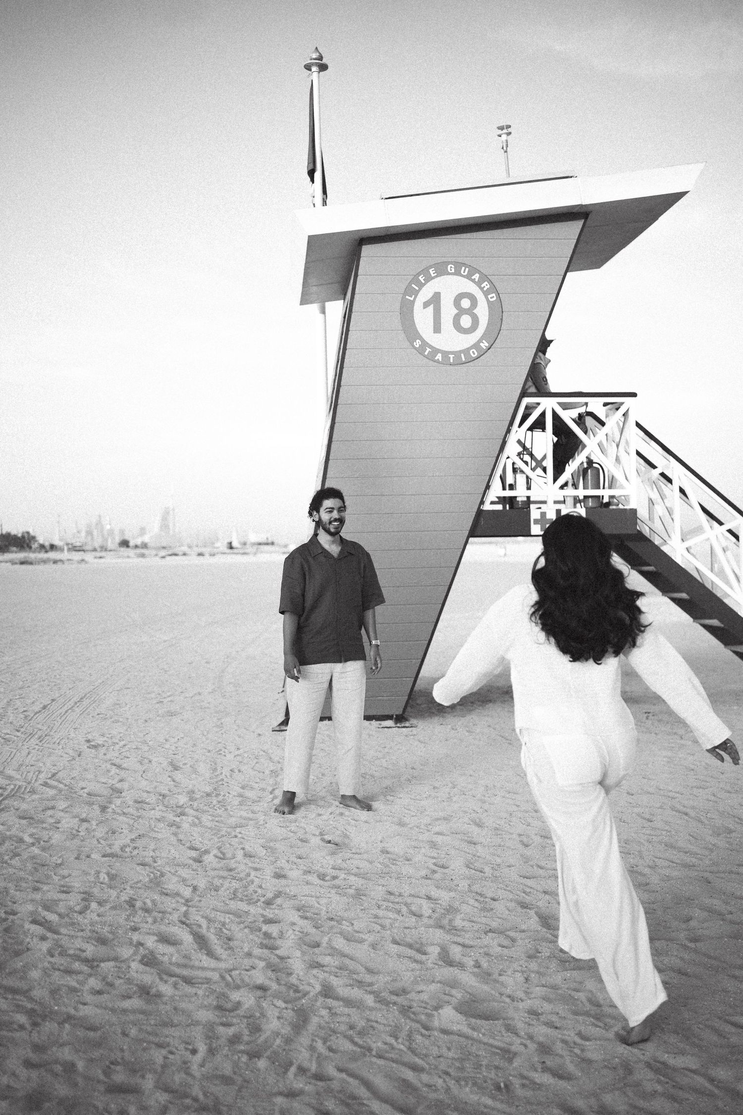 A couple poses playfully by lifeguard tower 18 on a beach in monochrome.