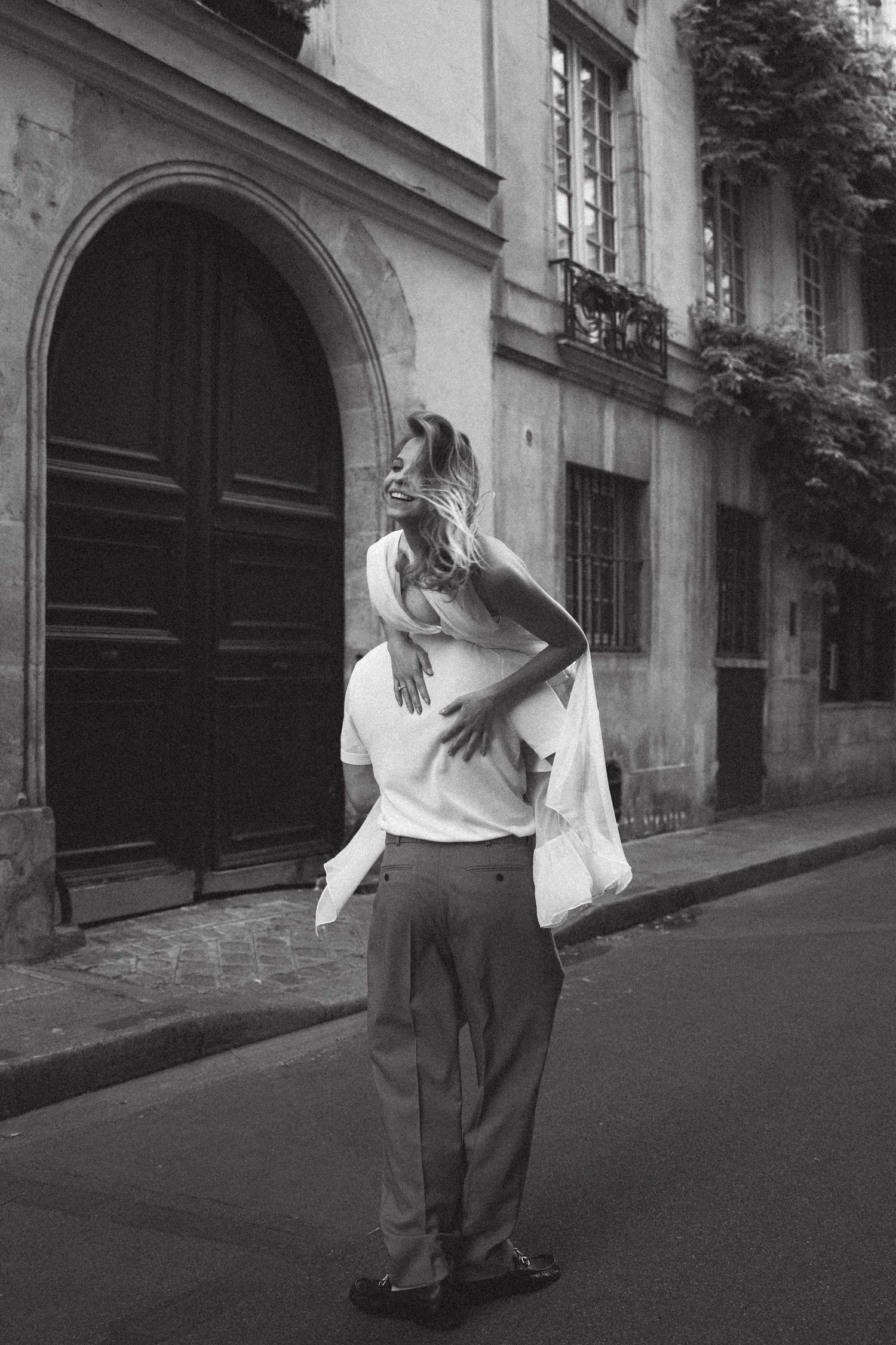 Black and white photo of a couple embracing on a Parisian street, with classic architecture in the background.