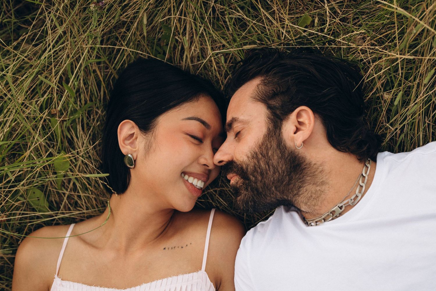 A smiling couple lying in green grass, sharing a romantic moment face to face outdoors.