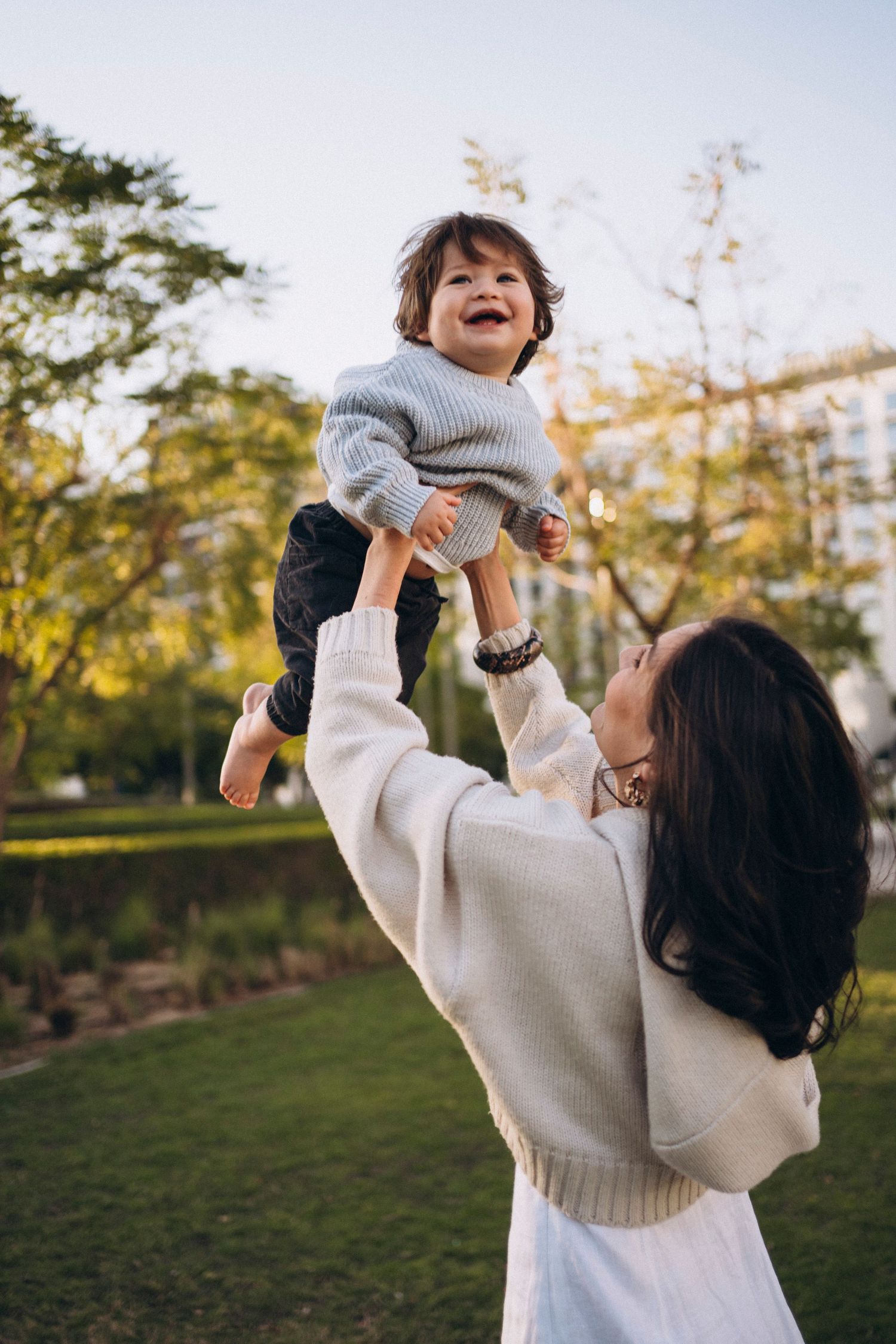 A parent lifts and plays with a small child wearing a grey sweater in a sunny outdoor garden setting.