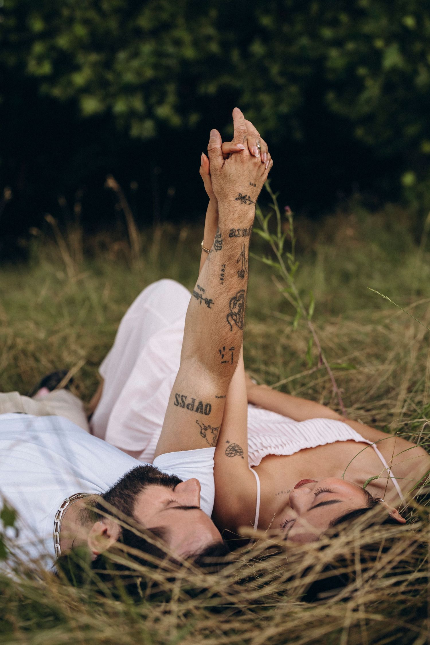 Couple lying in grass holding hands with tattooed arms raised, wearing white, eyes closed in a romantic outdoor setting.