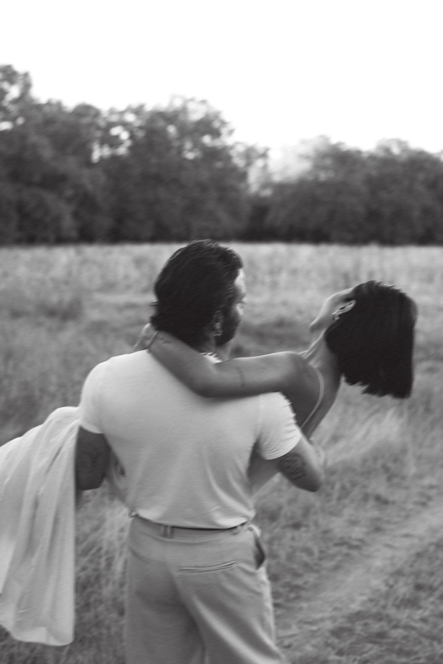 Black and white photo of a couple embracing in a field, man carrying woman in flowing dress near treeline.
