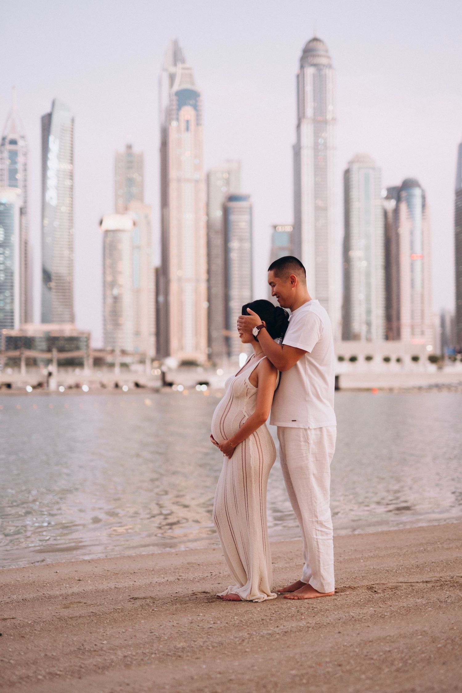 Couple embraces on beach with Dubai skyline in background, showcasing modern architecture and waterfront.