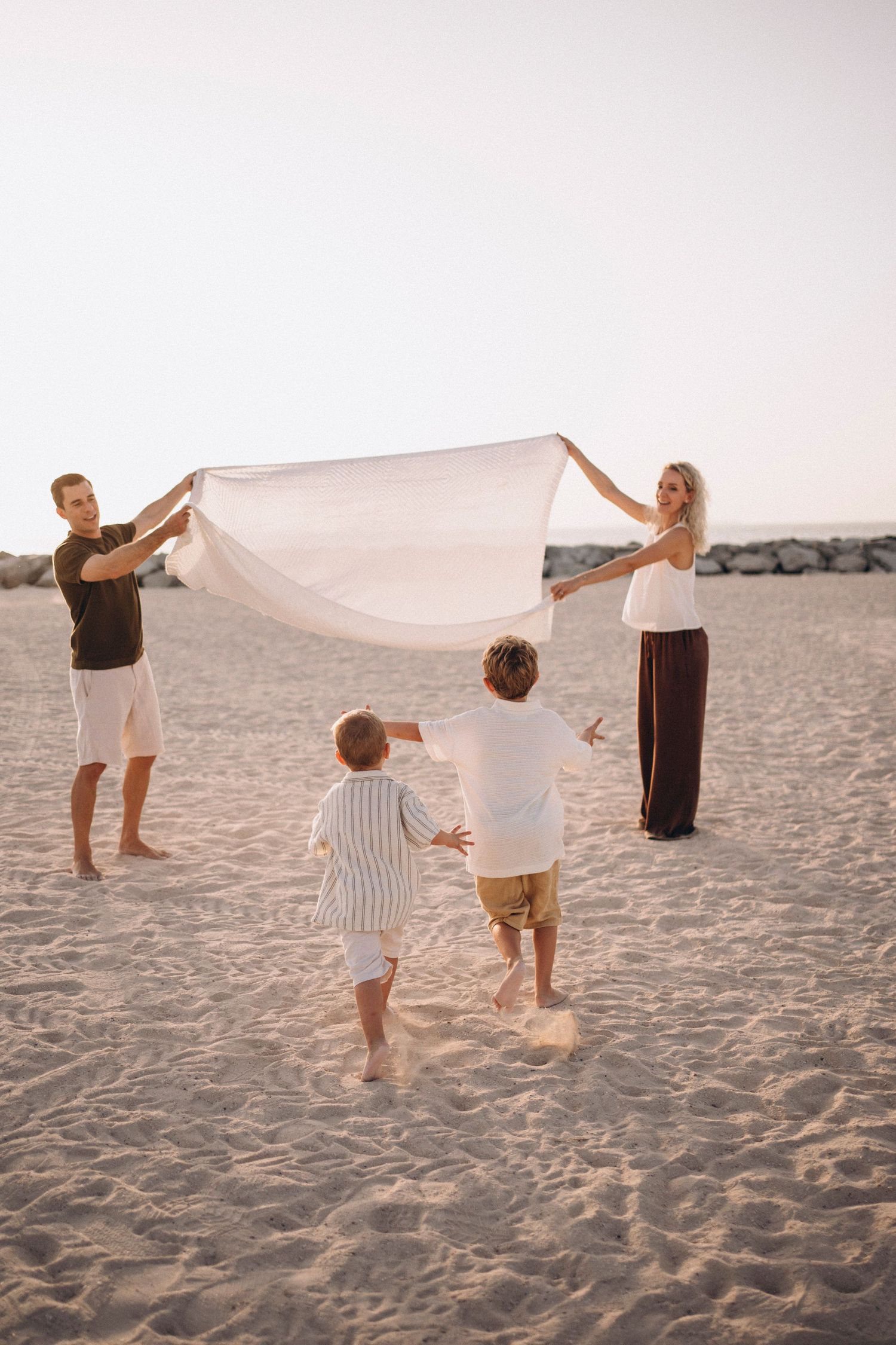 Family plays together on sandy beach at sunset holding white fabric in the breeze.