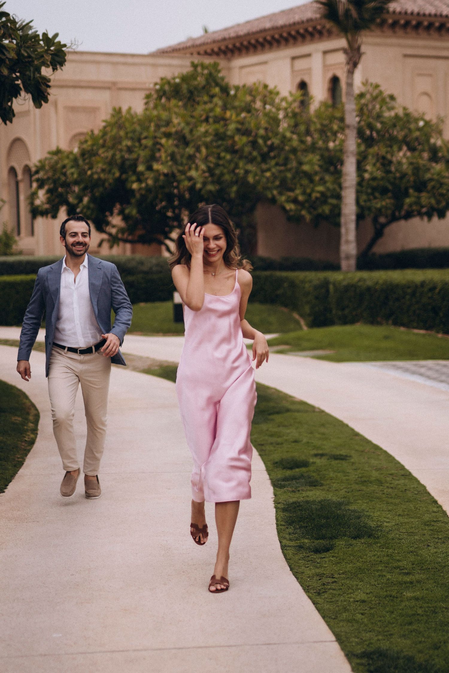 A couple shares a lighthearted moment walking along a manicured path by Spanish architecture.