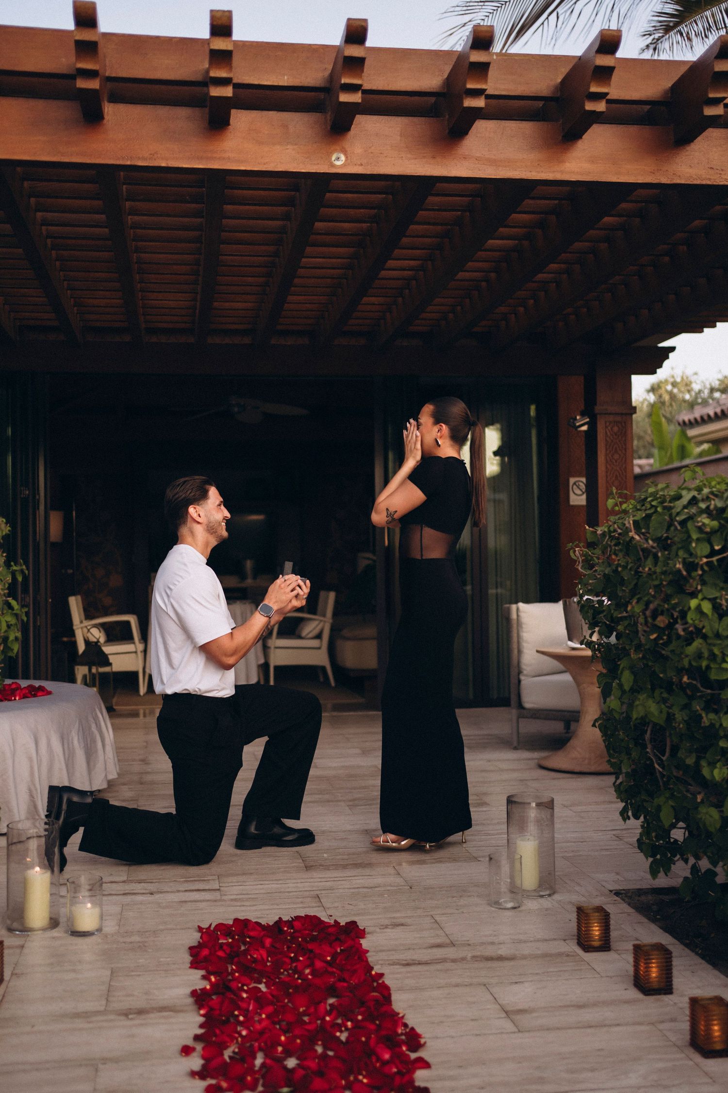 A romantic proposal setup on a patio with scattered red rose petals and glowing lanterns at sunset.