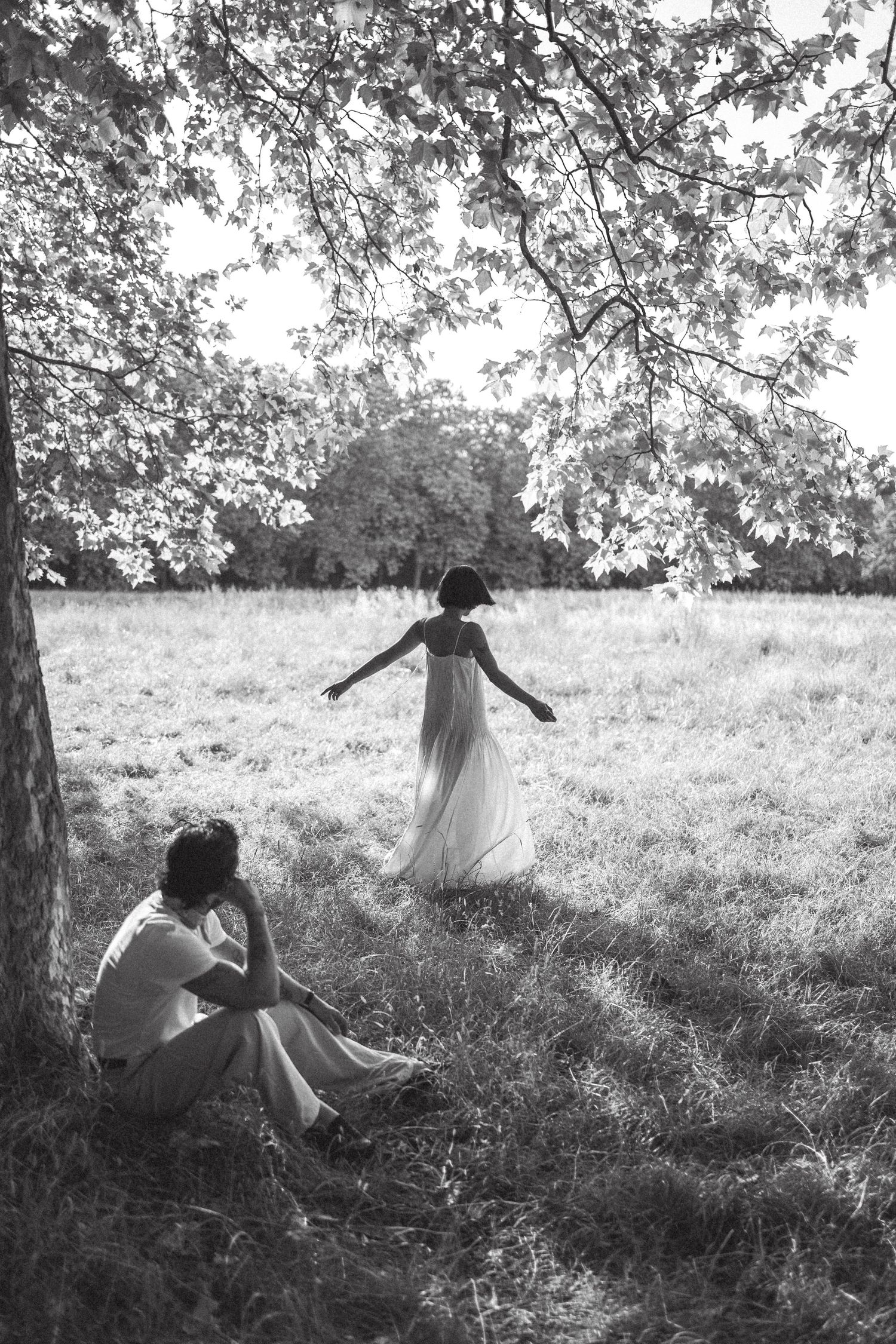 Black and white photo of two women in white dresses in a field, one sitting under a tree, one walking away.