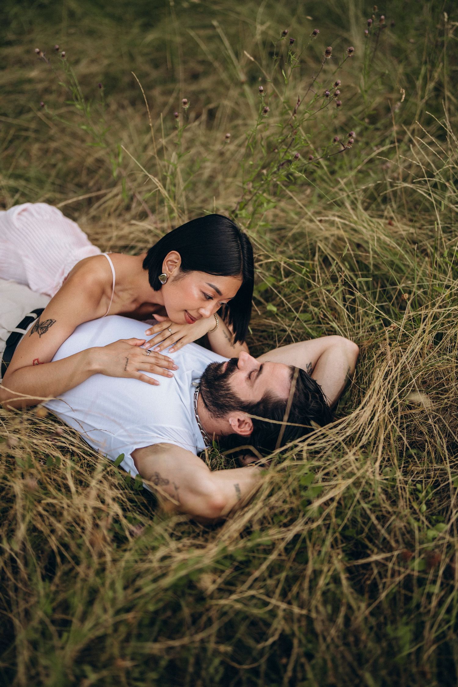 A romantic couple lying in tall green grass, sharing a tender kiss in a serene outdoor meadow setting.