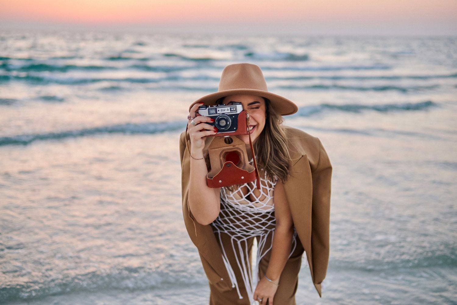 A photographer in a brown hat takes pictures at the beach during golden hour.