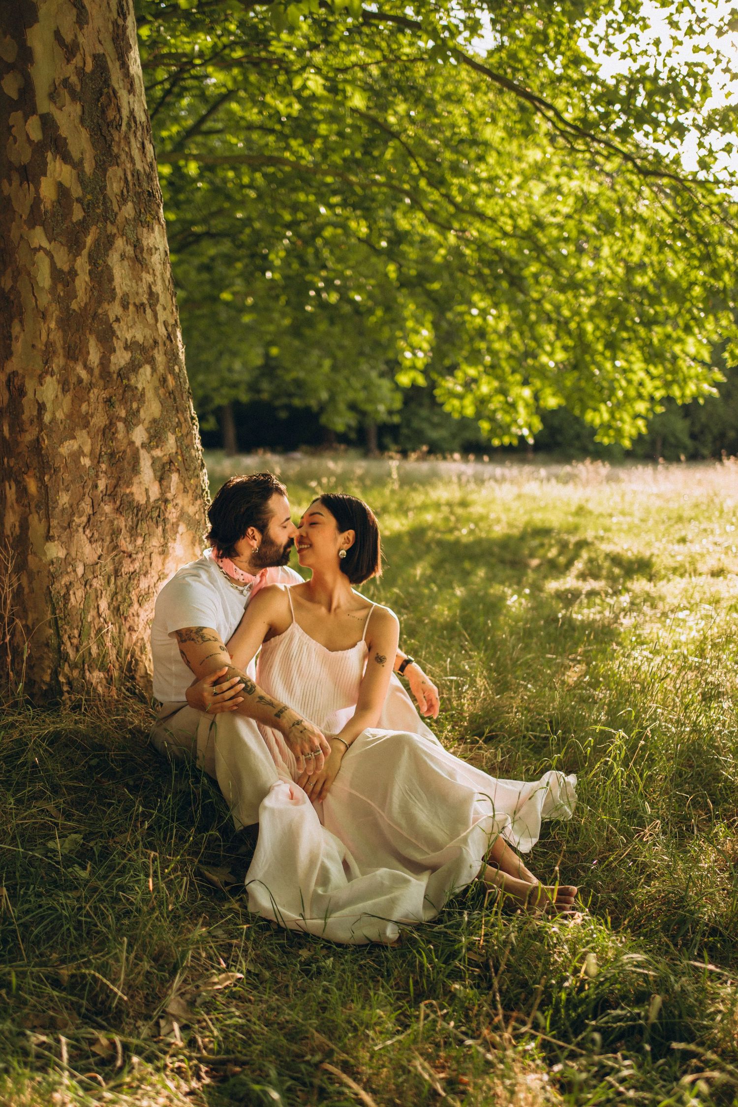 Couple sitting against a tree in a sunny green park, sharing a romantic moment in golden afternoon light.