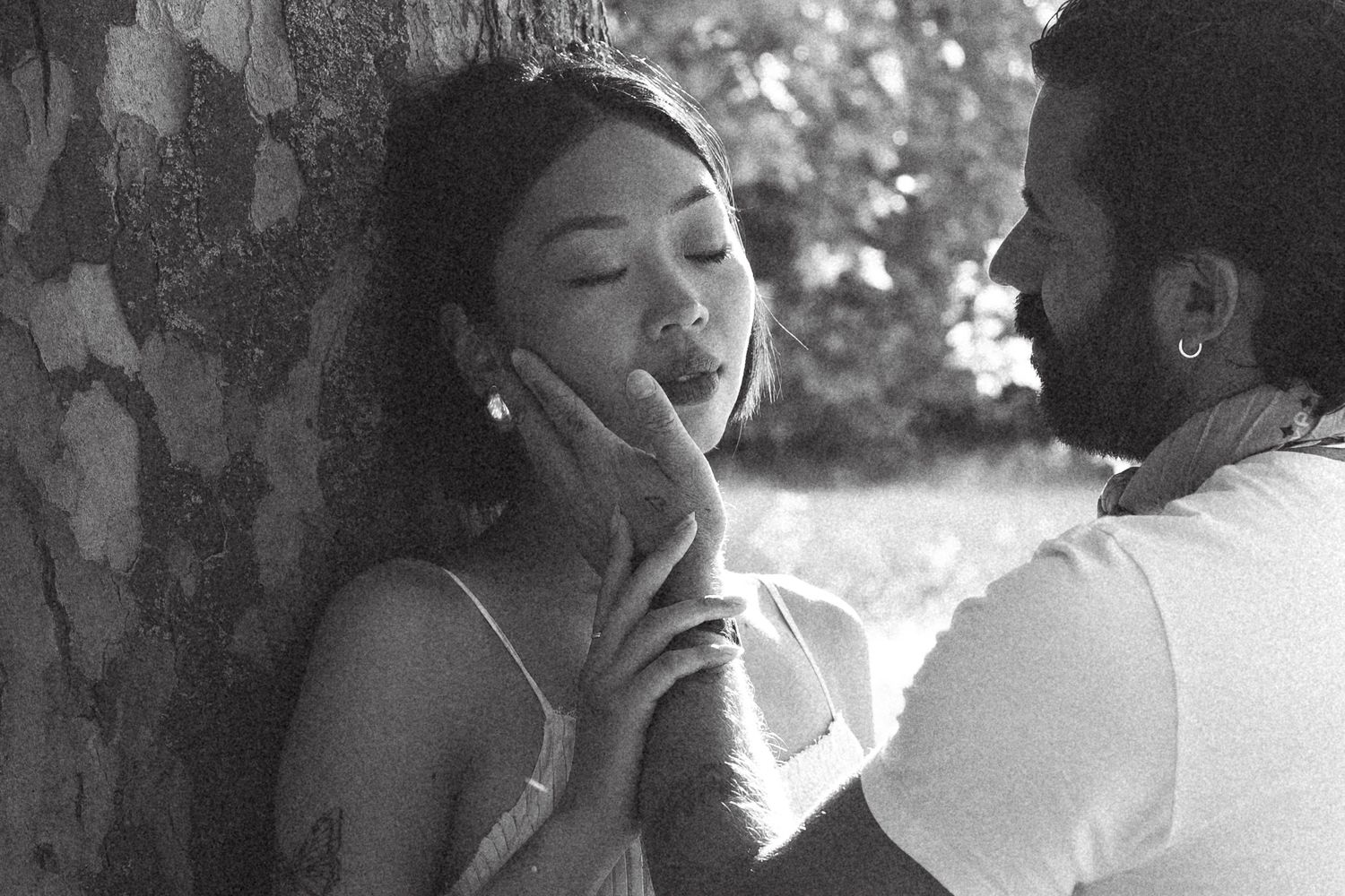 Black and white romantic photo of a couple sharing an intimate moment outdoors near a tree.