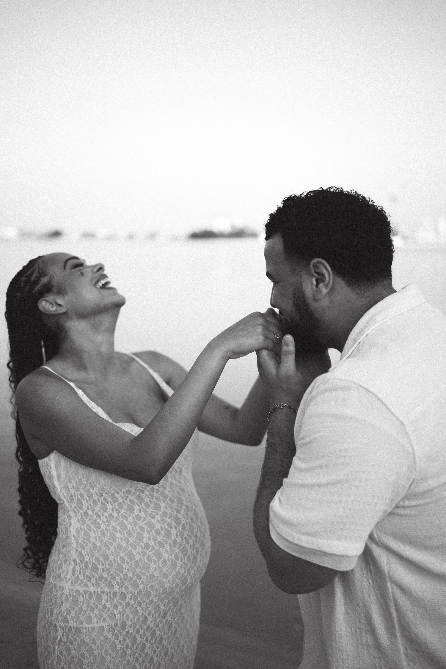 Black and white photo of expecting couple sharing playful moment on beach.