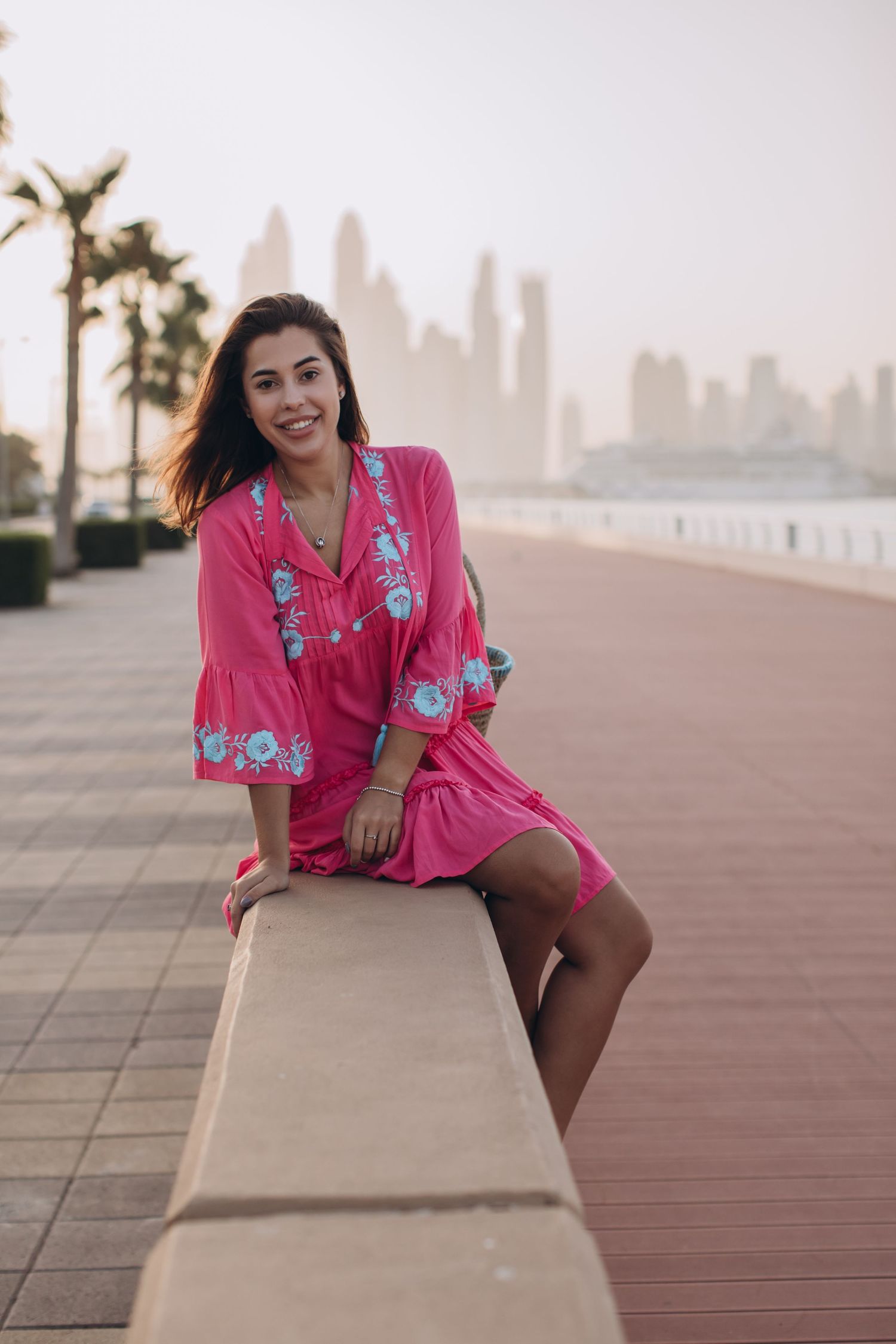 A person in a pink floral dress sits on a concrete wall with Dubai's skyline and palm trees in the background.