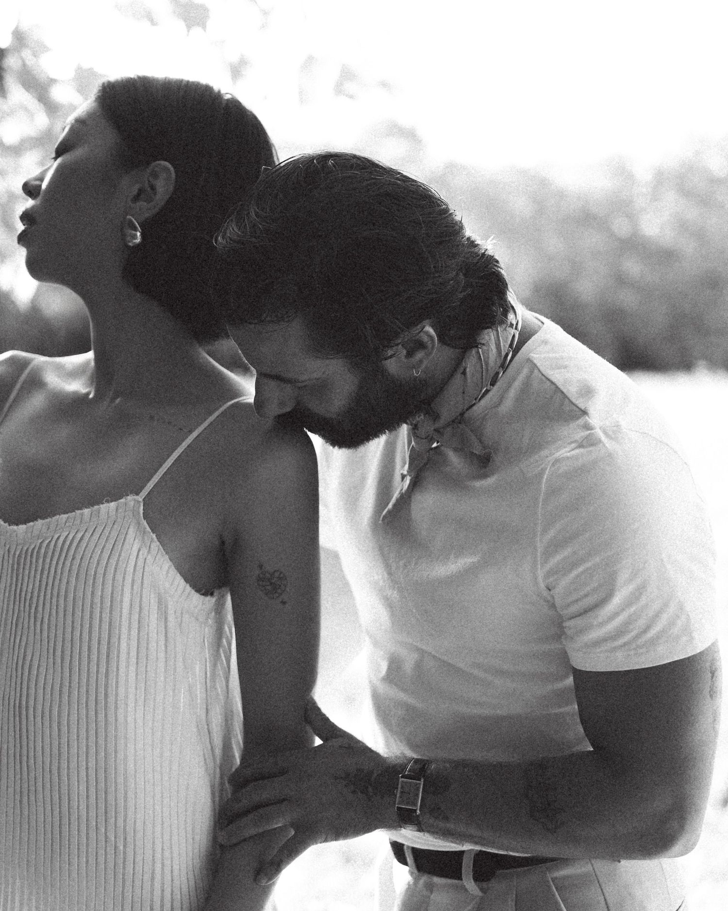 Black and white photo of a couple in an intimate embrace outdoors, man kissing woman's shoulder in romantic pose.