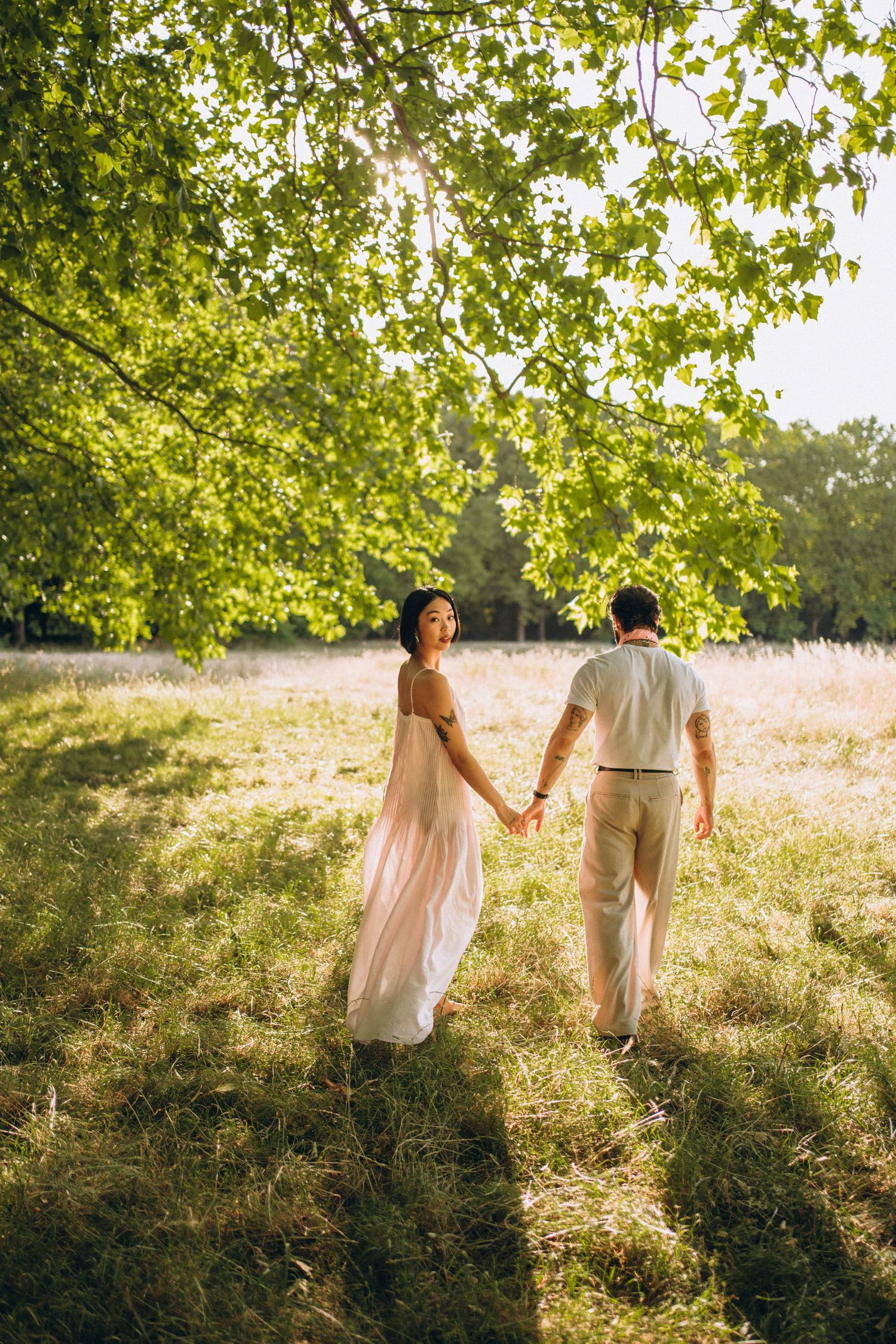 Couple holding hands walking through sunlit meadow, woman in flowing pink dress, man in cream outfit, lush green tree overhead.