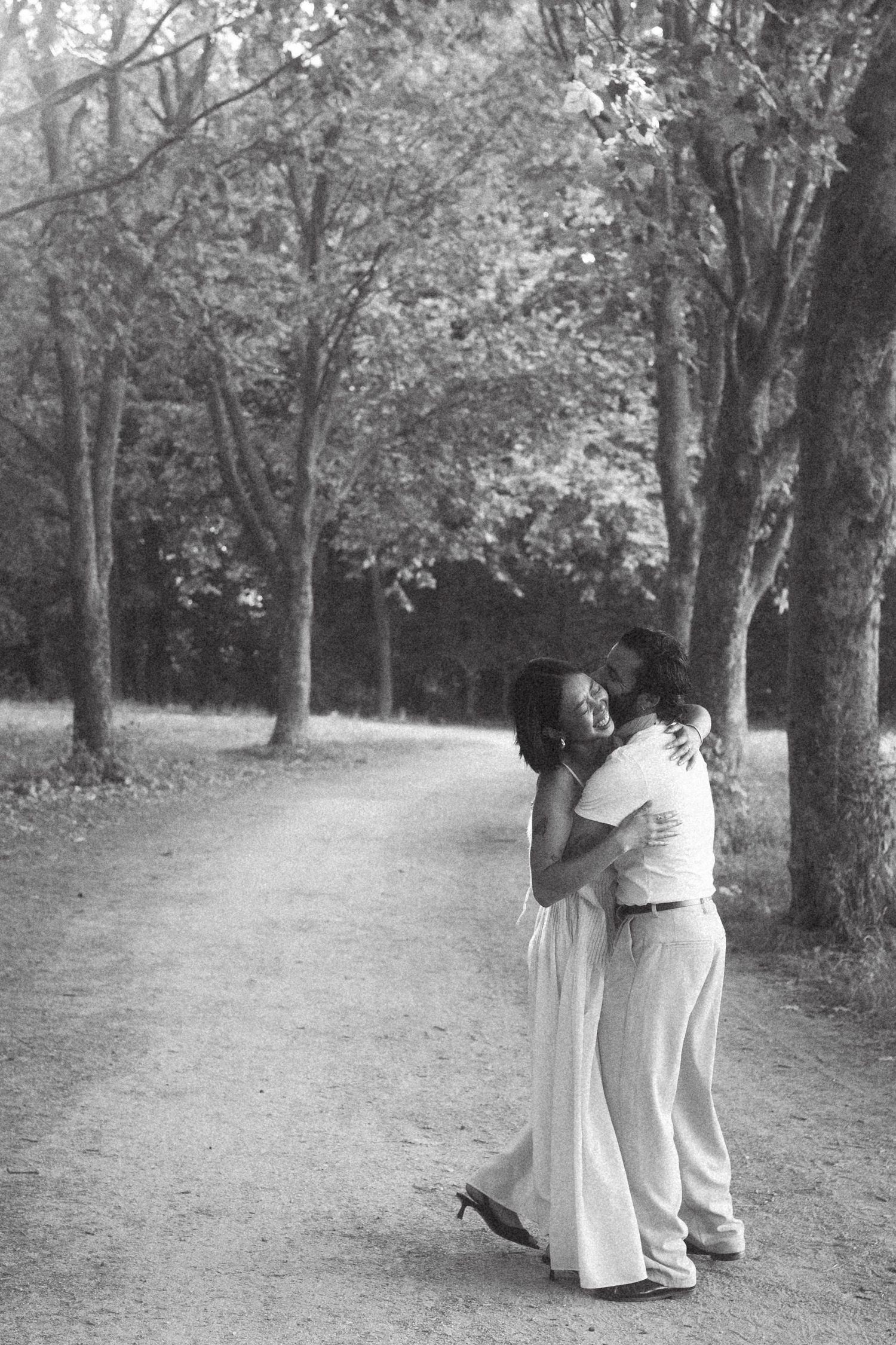 Black and white photo of a couple embracing and smiling on a tree-lined path in a park.