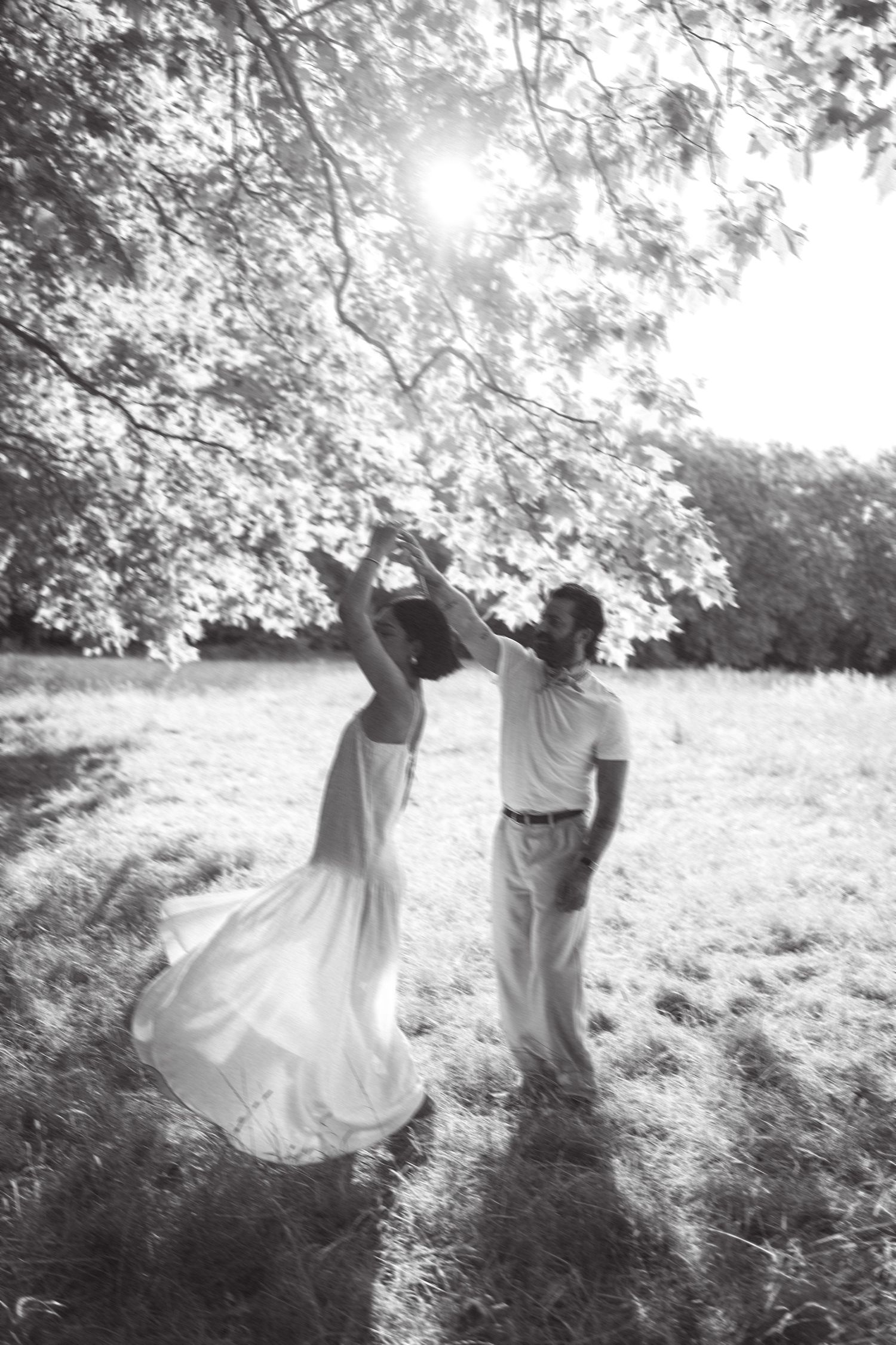 Black and white photo of a couple dancing outdoors under a tree, woman in flowing white dress being twirled by man.