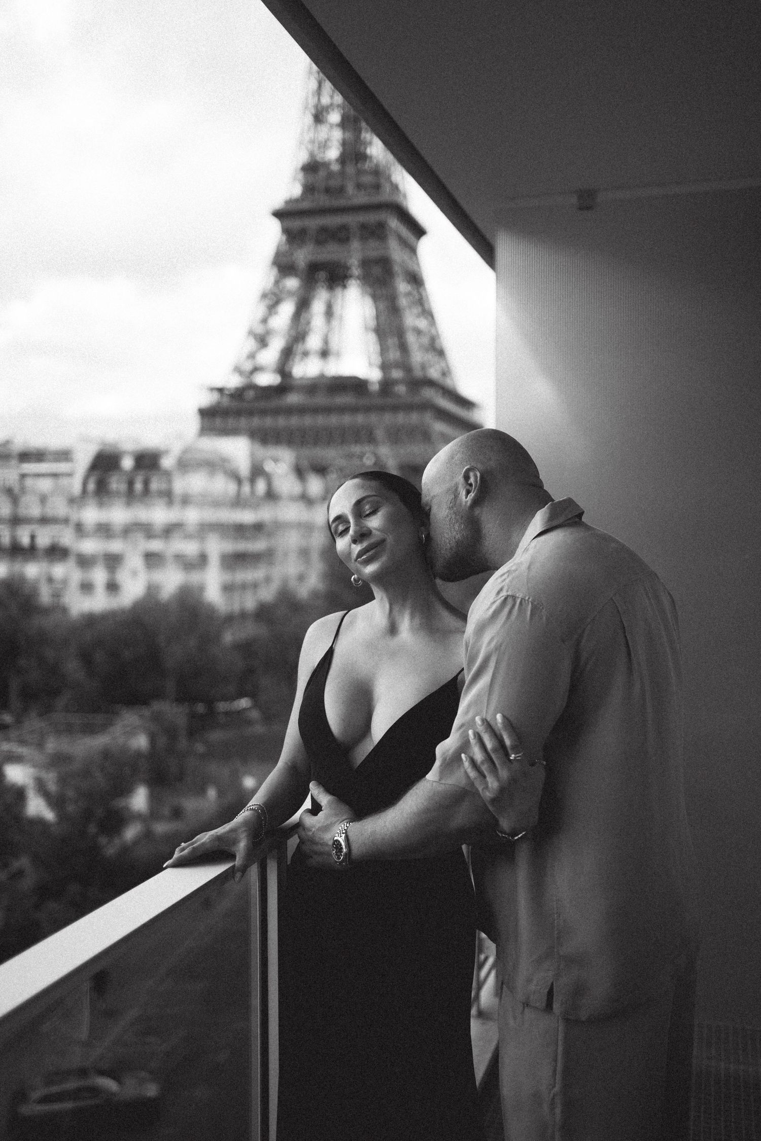 Black and white photo of a romantic couple embracing on a balcony with the Eiffel Tower in Paris behind them.