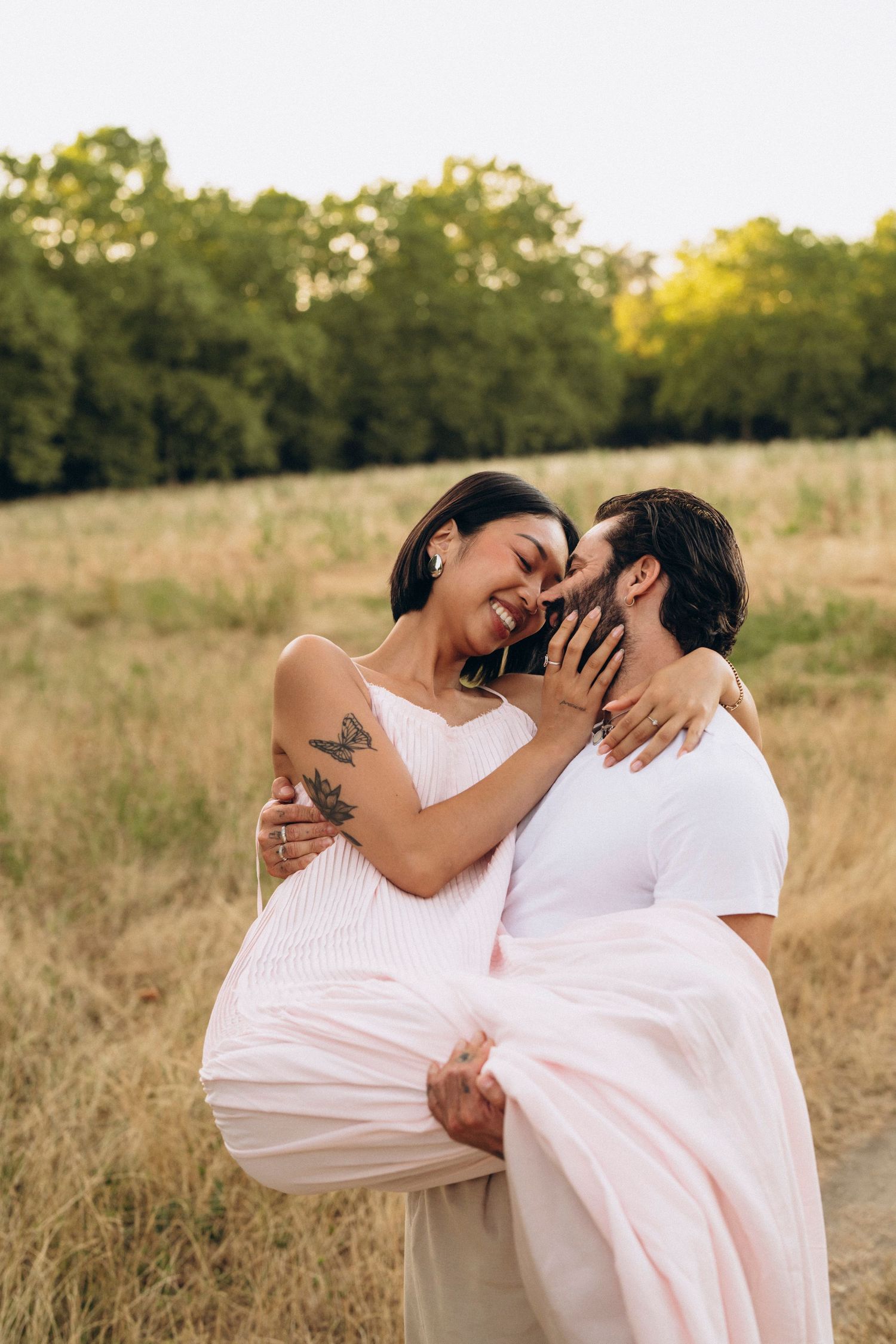 A man carries a laughing woman in a pink dress outdoors in a golden field, sharing a romantic moment.