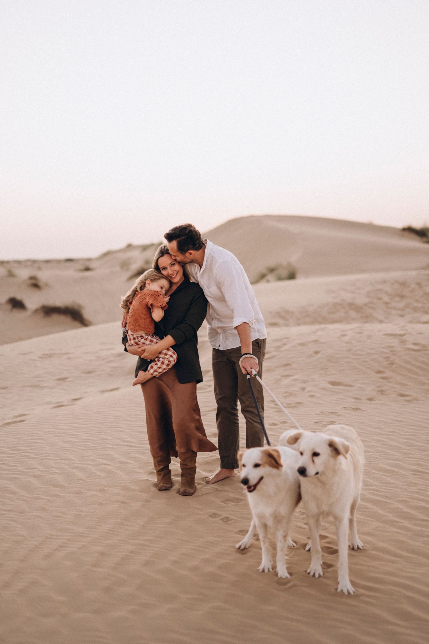 A tender family moment captured on sandy dunes with two white dogs in the foreground.