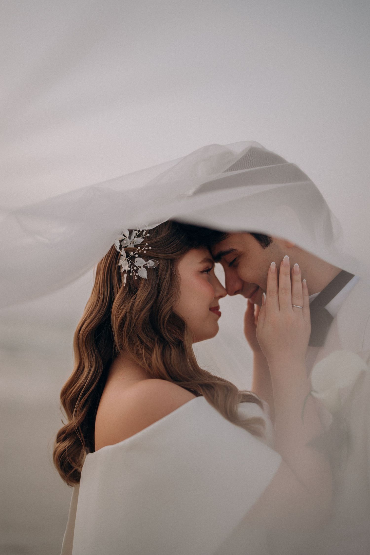 Bride and groom share a romantic moment under a flowing white veil, with the bride wearing a floral hair accessory.