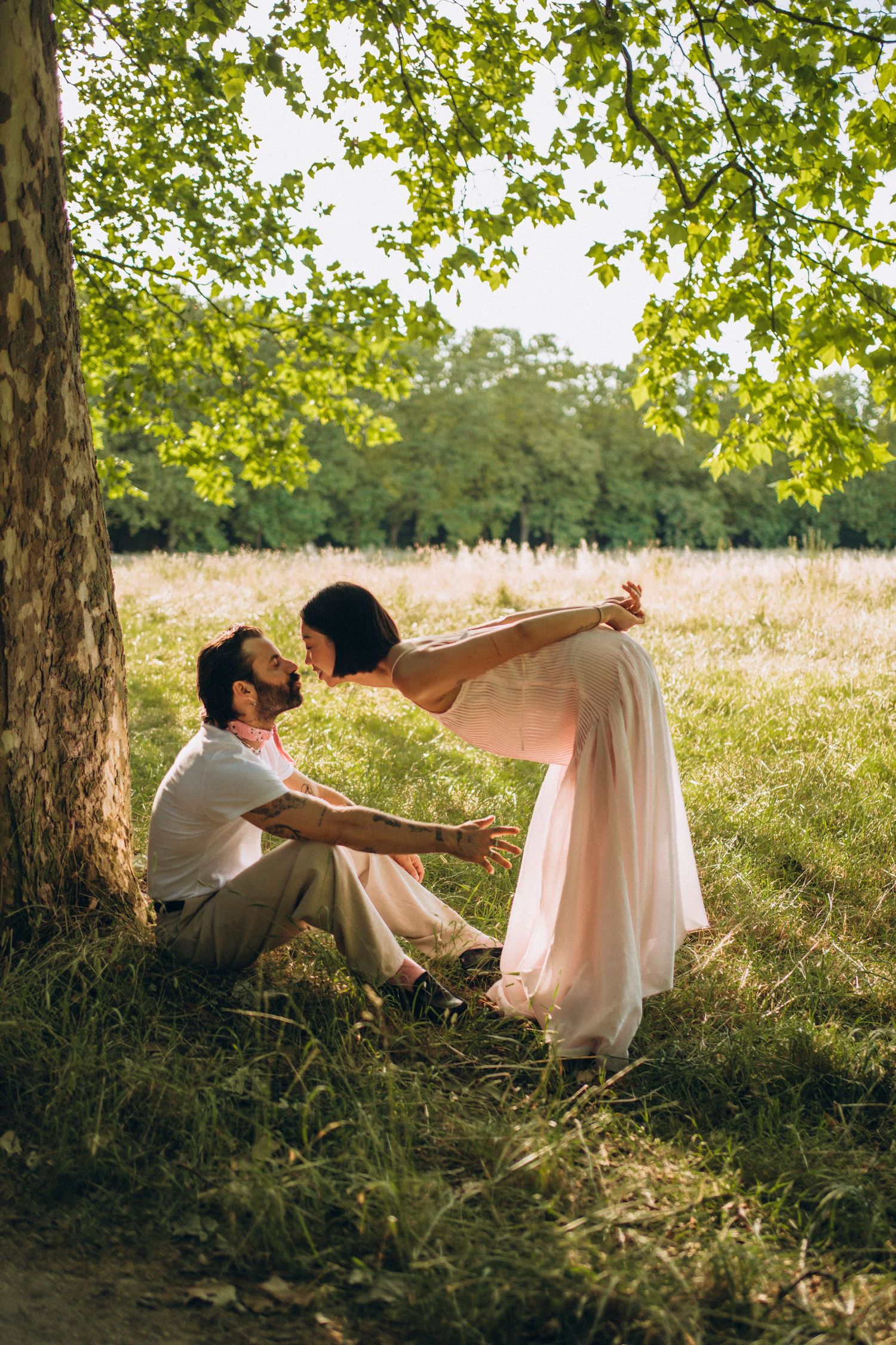 Romantic couple sitting under a tree in a sunny meadow, woman in pink dress leaning toward man in white shirt.