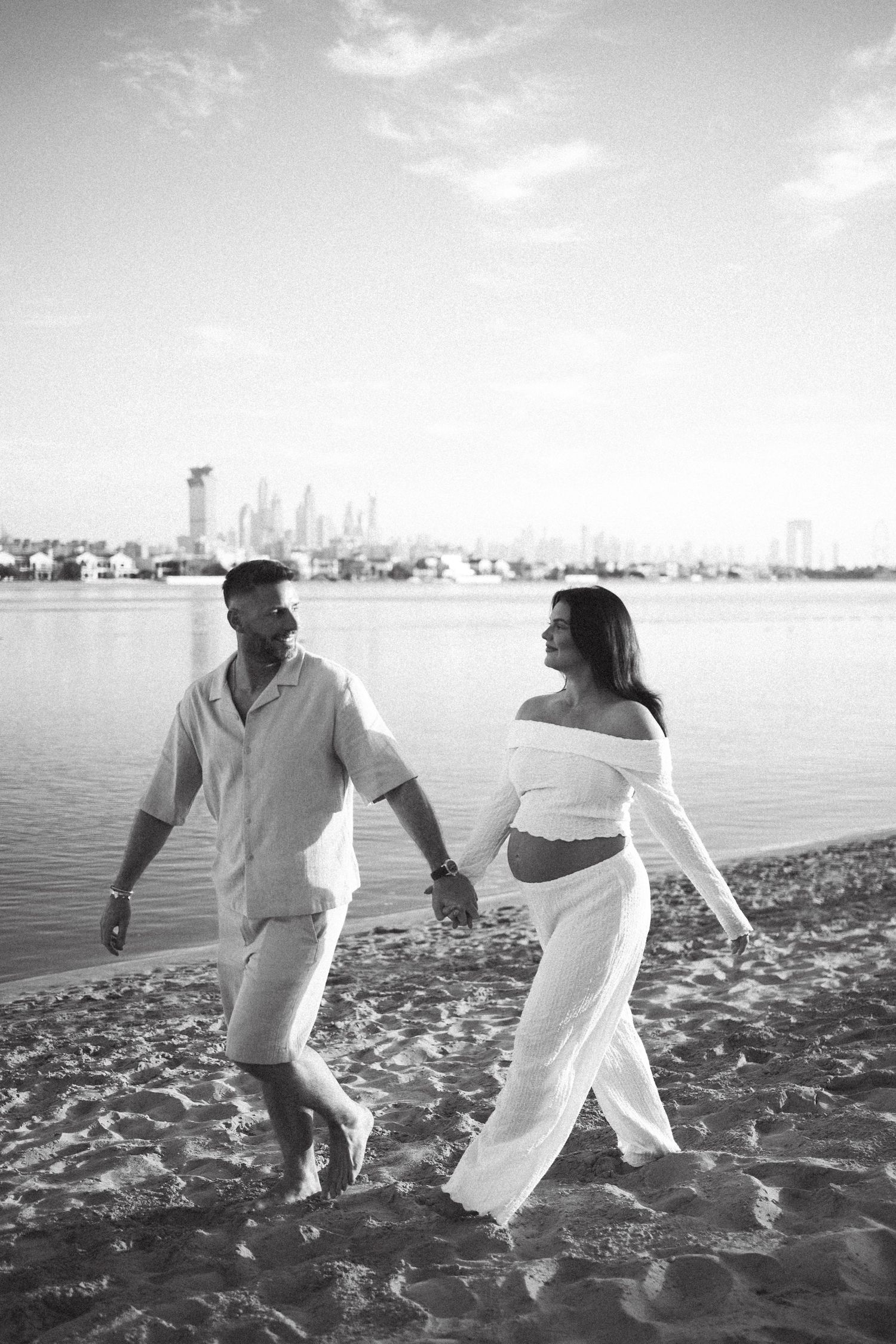 A couple in white outfits walks along a beach with a city skyline in the background.