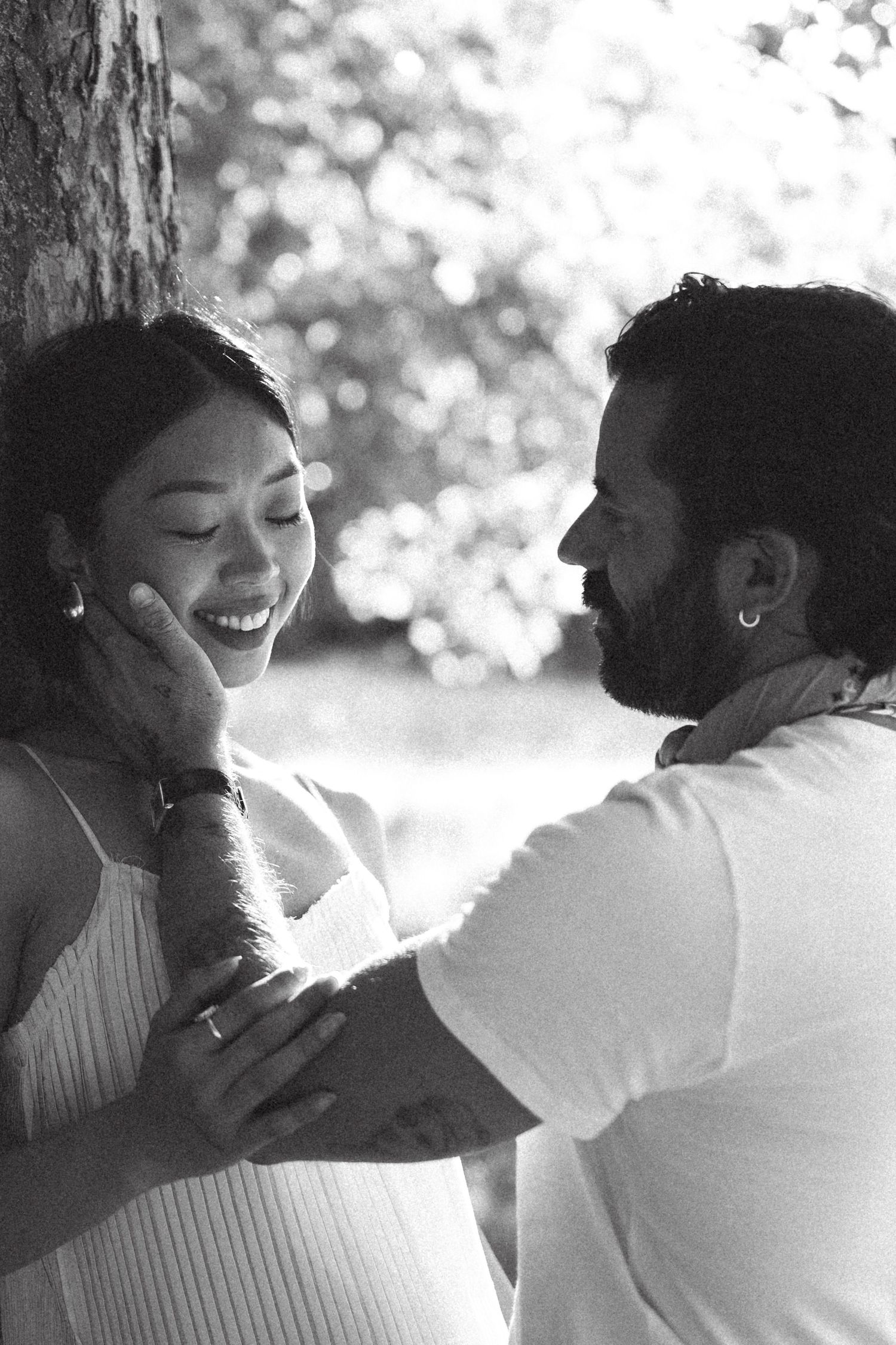 Black and white photo of a smiling couple sharing a tender moment, man gently holding womans face near a tree.