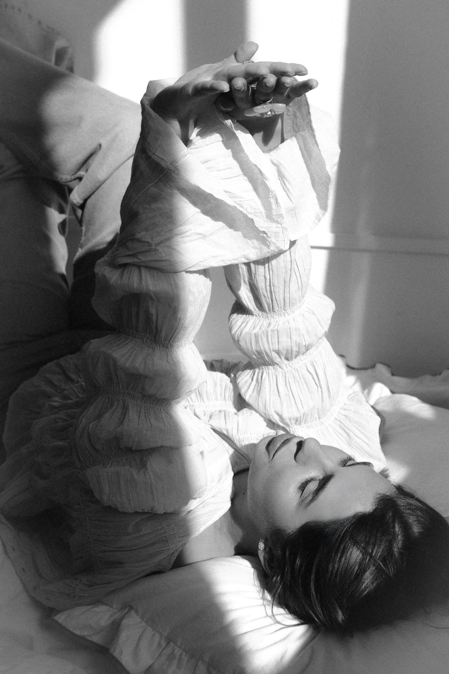 Black and white photo of a woman lying down in a white ruffled dress, holding a phone above her face in soft sunlight.