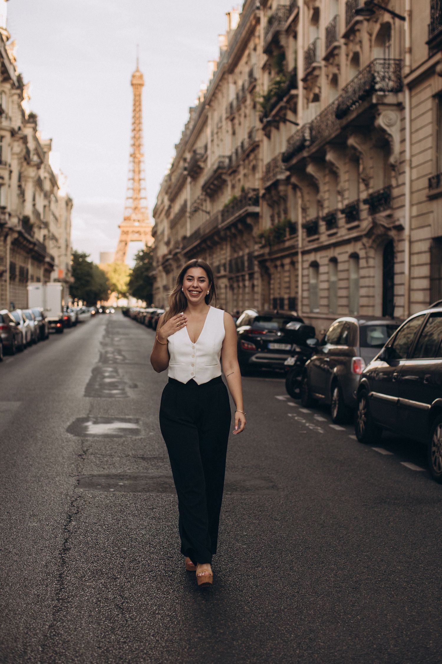 A woman walks down a Parisian street with the Eiffel Tower visible in the background at sunset.