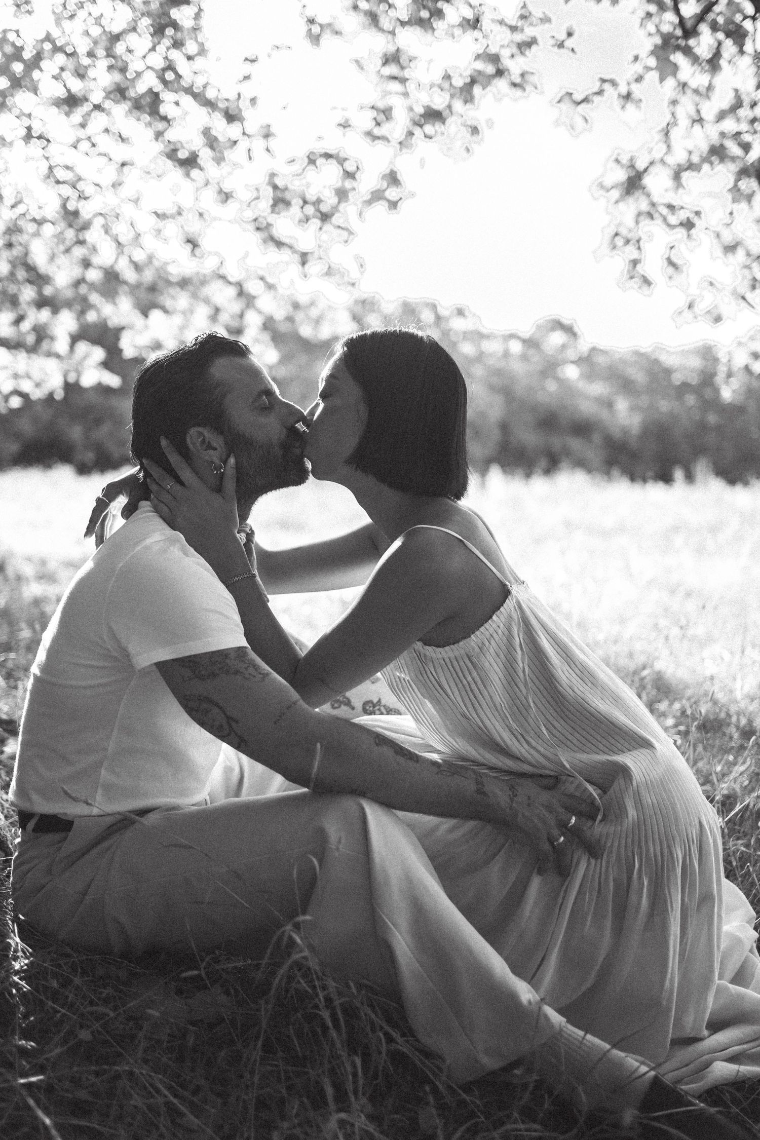 Black and white photo of a couple kissing while sitting in a sunny field beneath blooming trees.