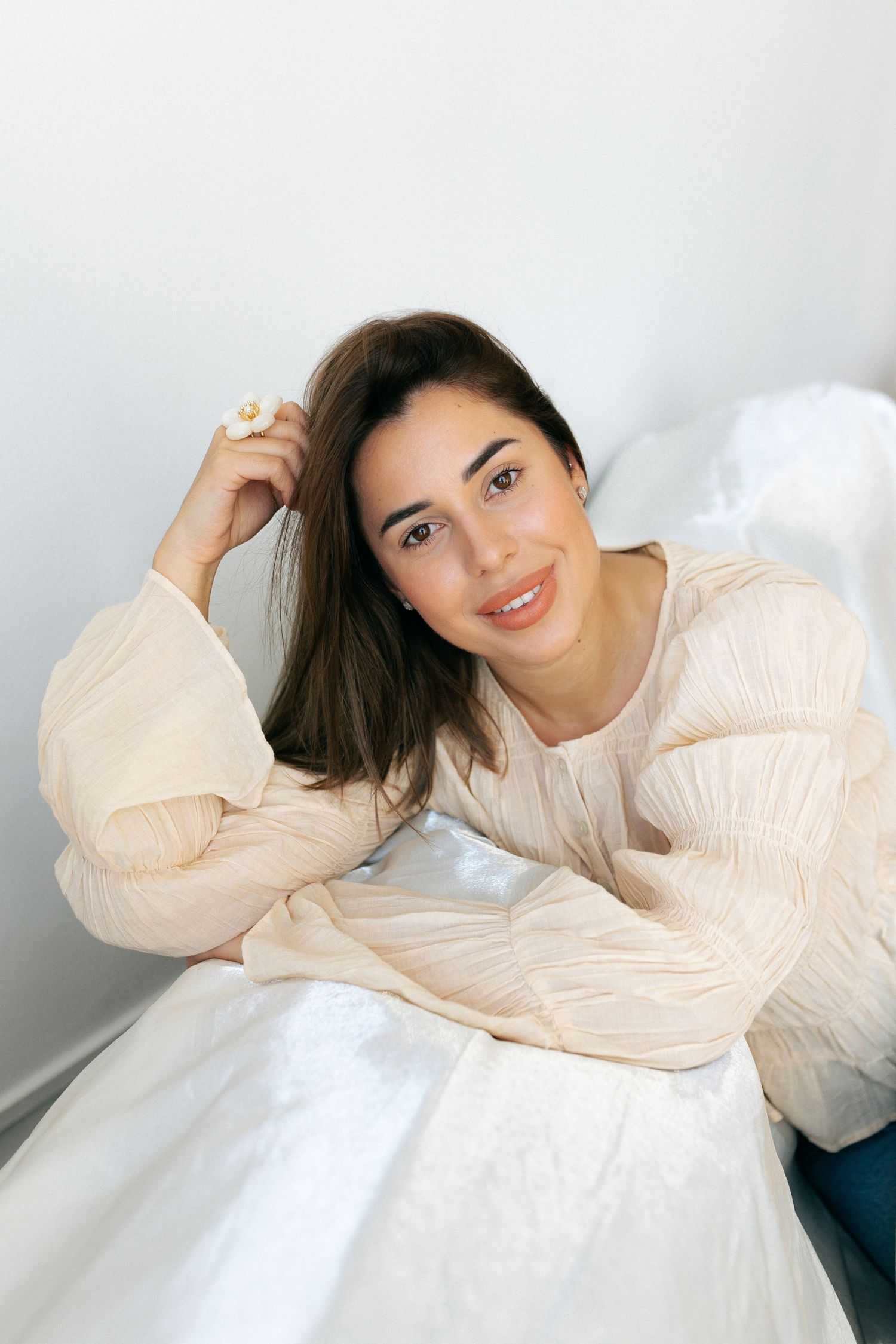 Young woman in cream blouse leaning on bed, smiling softly with hand in dark hair against white wall.