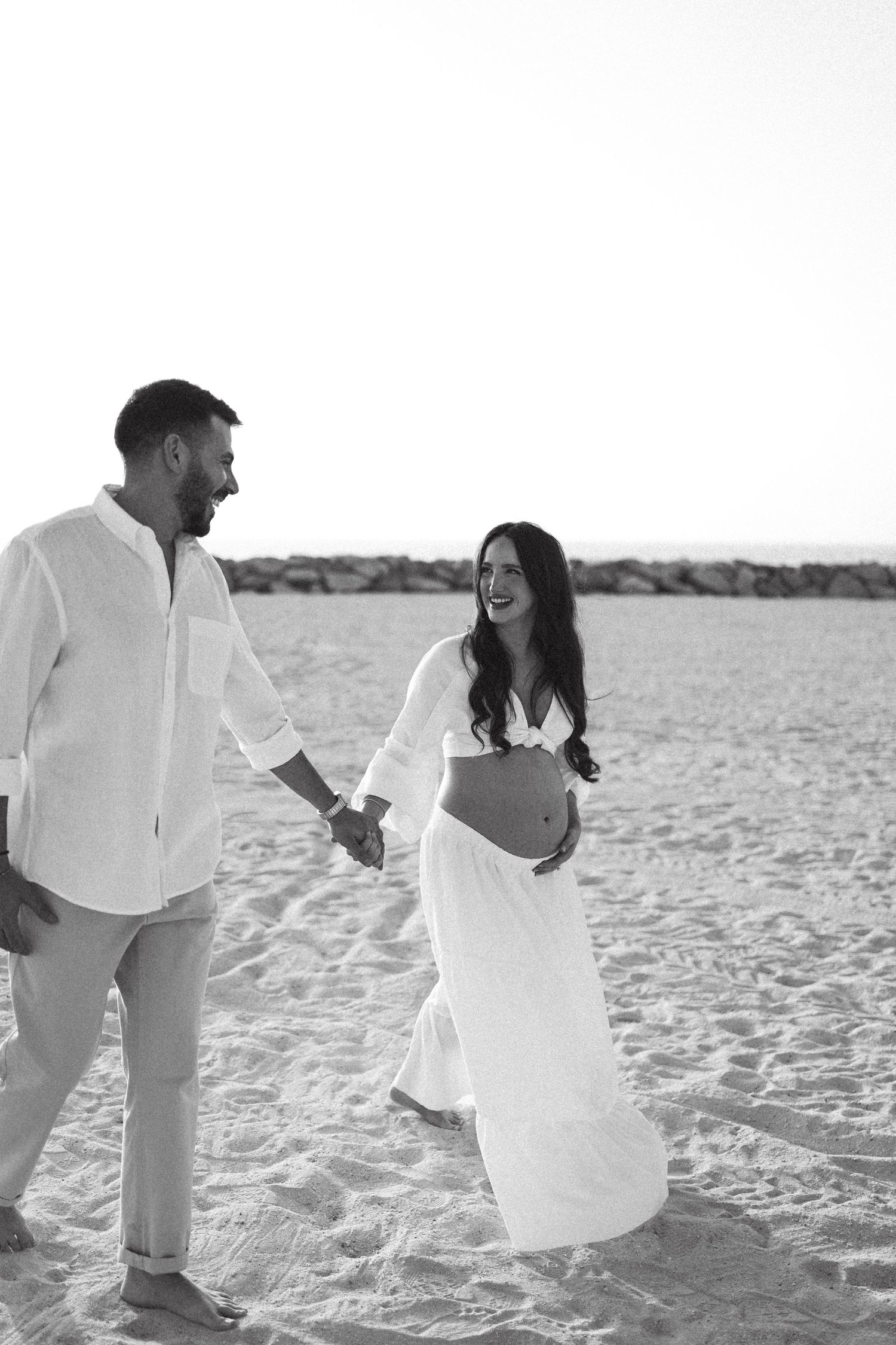 A couple playfully interacting on a bright sandy beach in white outfits.