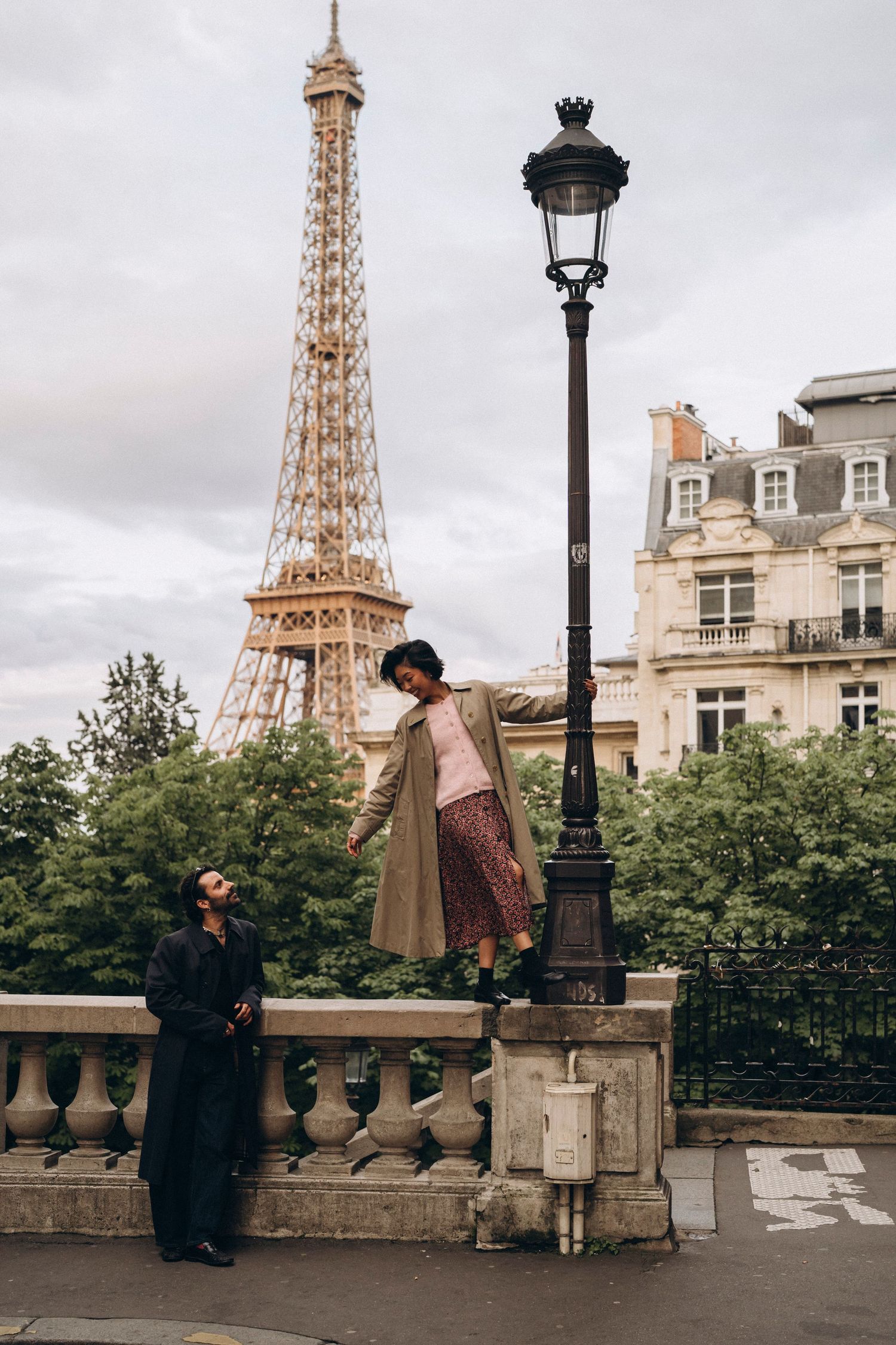 Couple posing near Eiffel Tower in Paris, woman standing on ledge by lamppost, man looking up, cloudy sky.