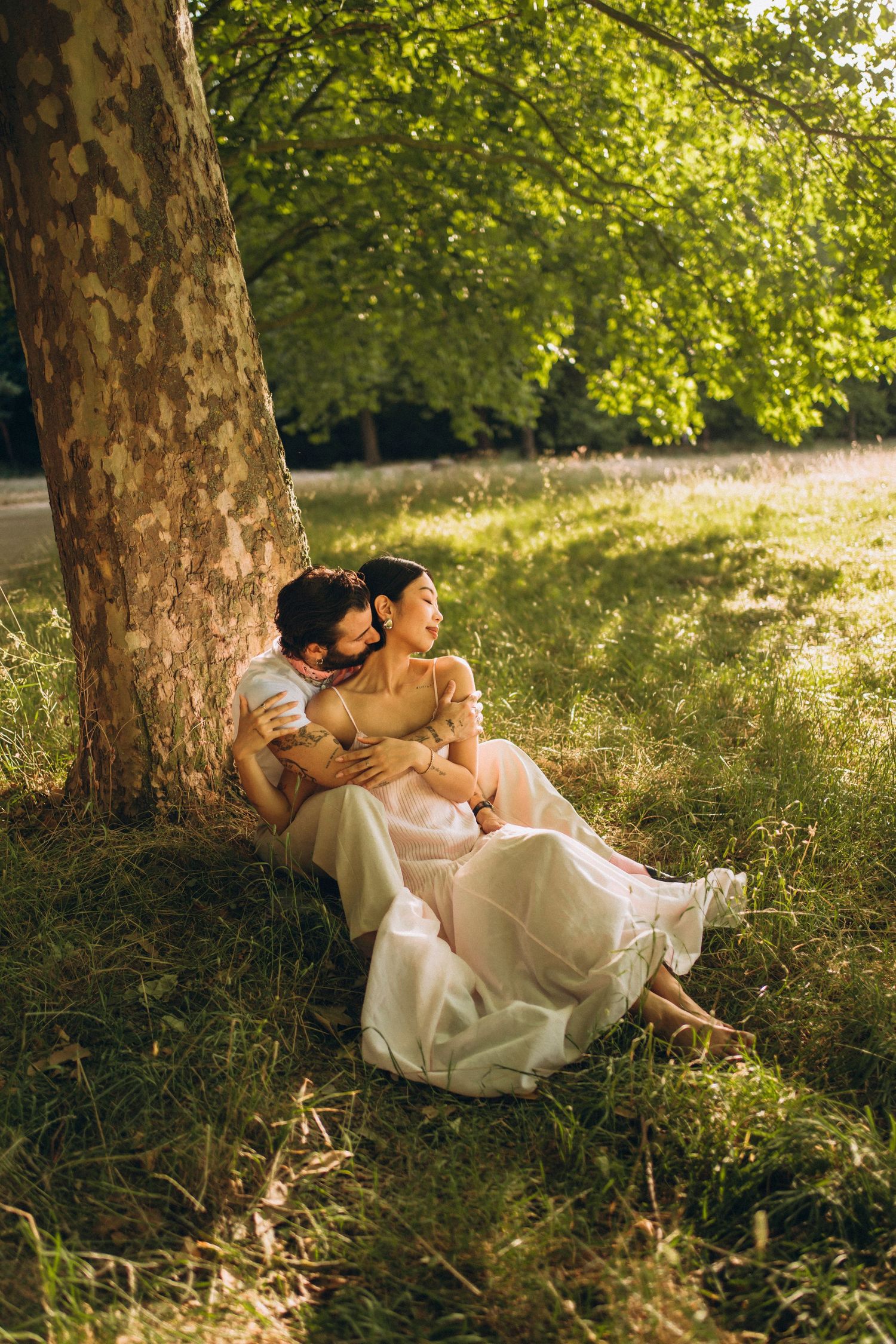 Couple sitting together against a tree trunk in a sunlit park, woman in flowing dress embraced by partner on lush grass.