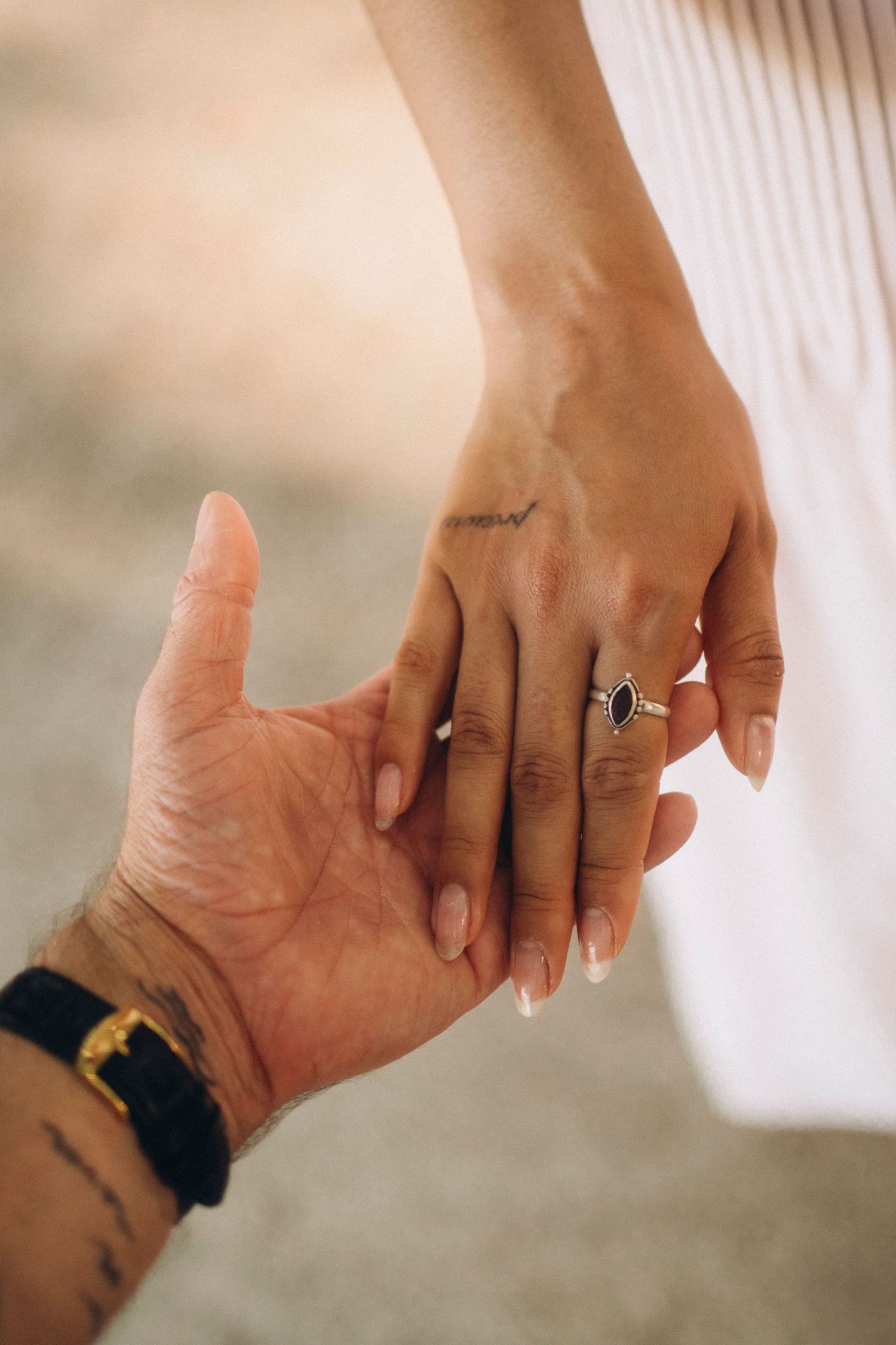 Close-up of two hands touching, woman wearing a black gemstone ring and small tattoo, man with bracelet and tattoo.