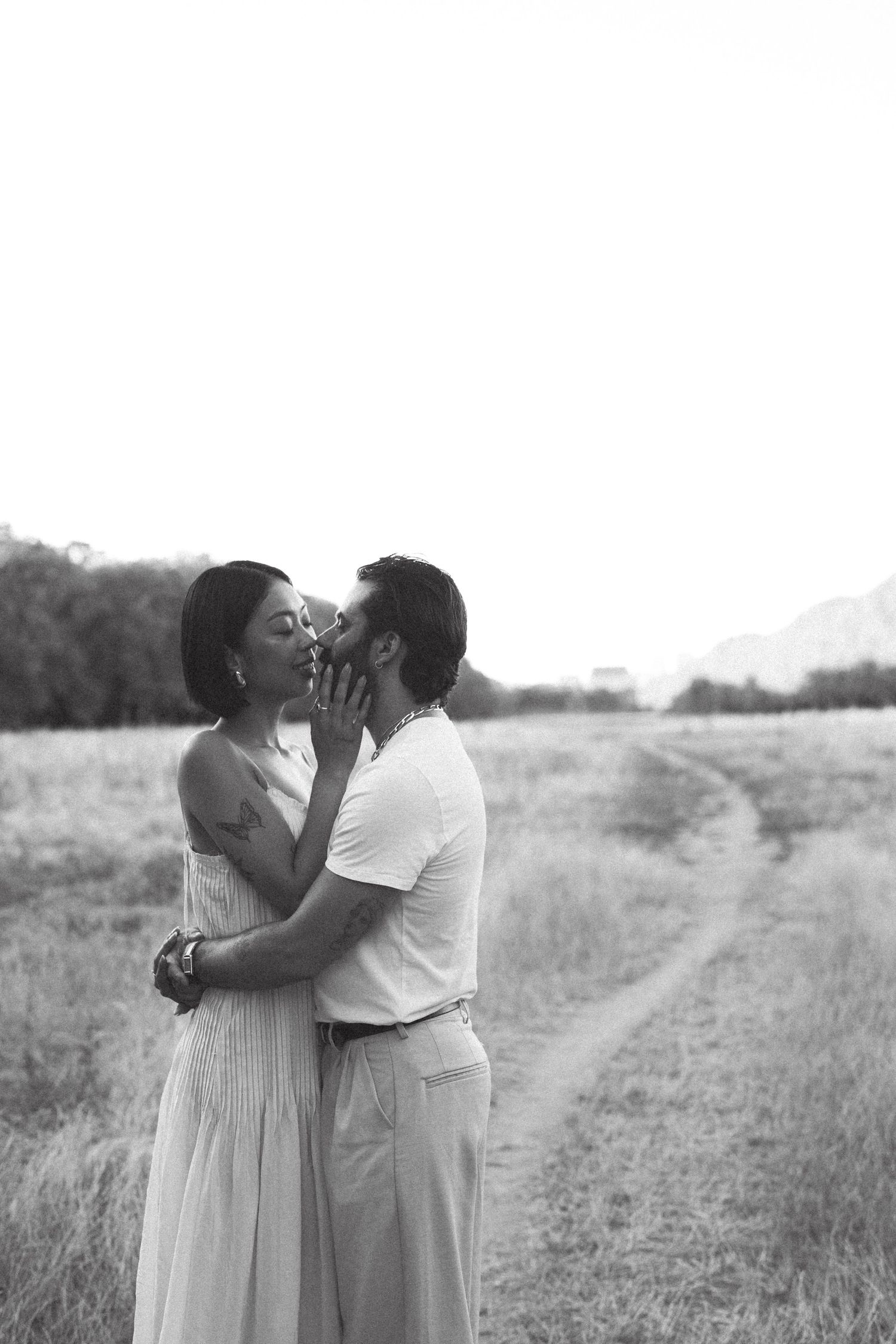 Black and white photo of a romantic couple kissing in a scenic field with a winding path and mountains in the background.