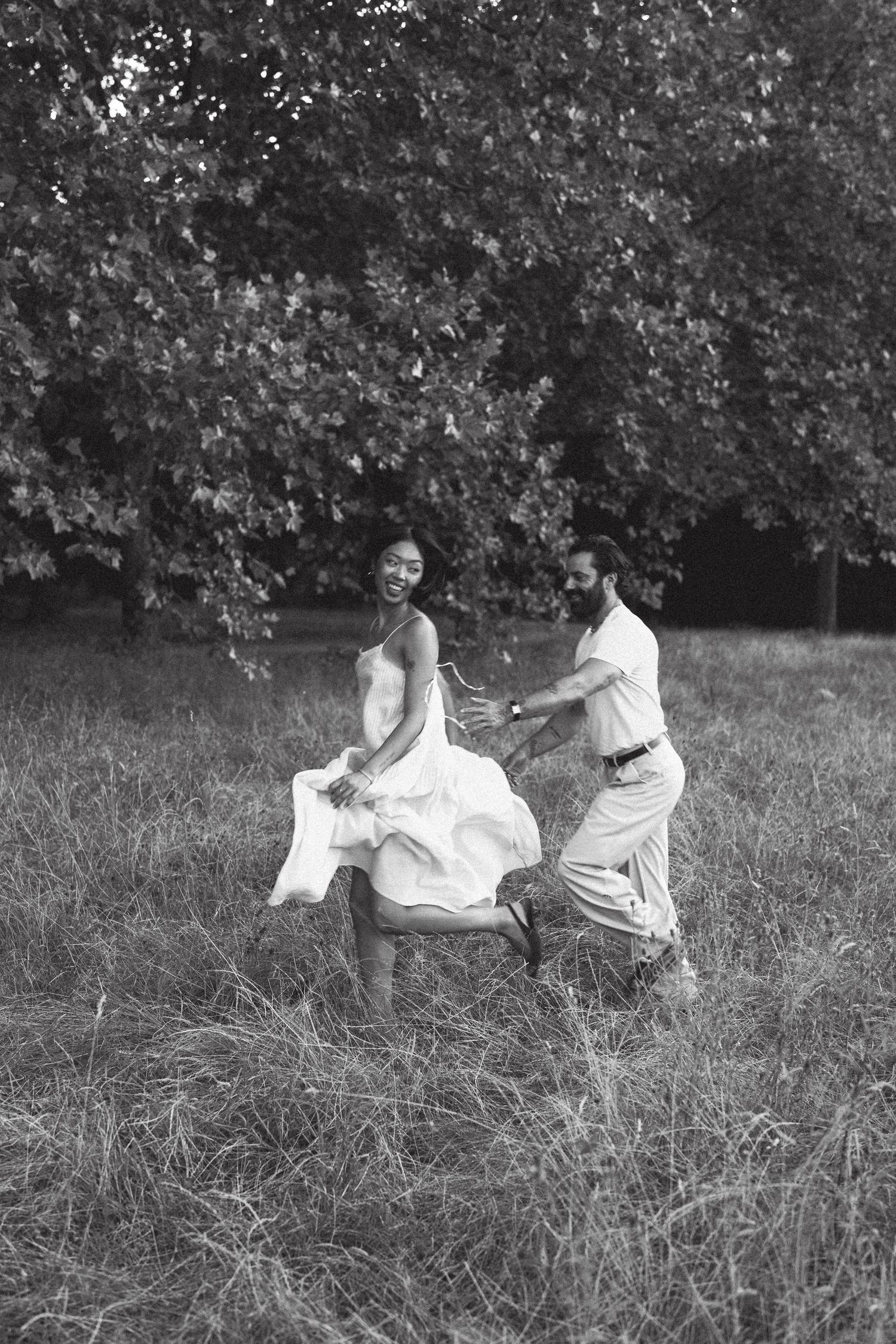 Black and white photo of a joyful couple dancing in a grassy field with lush trees in the background.