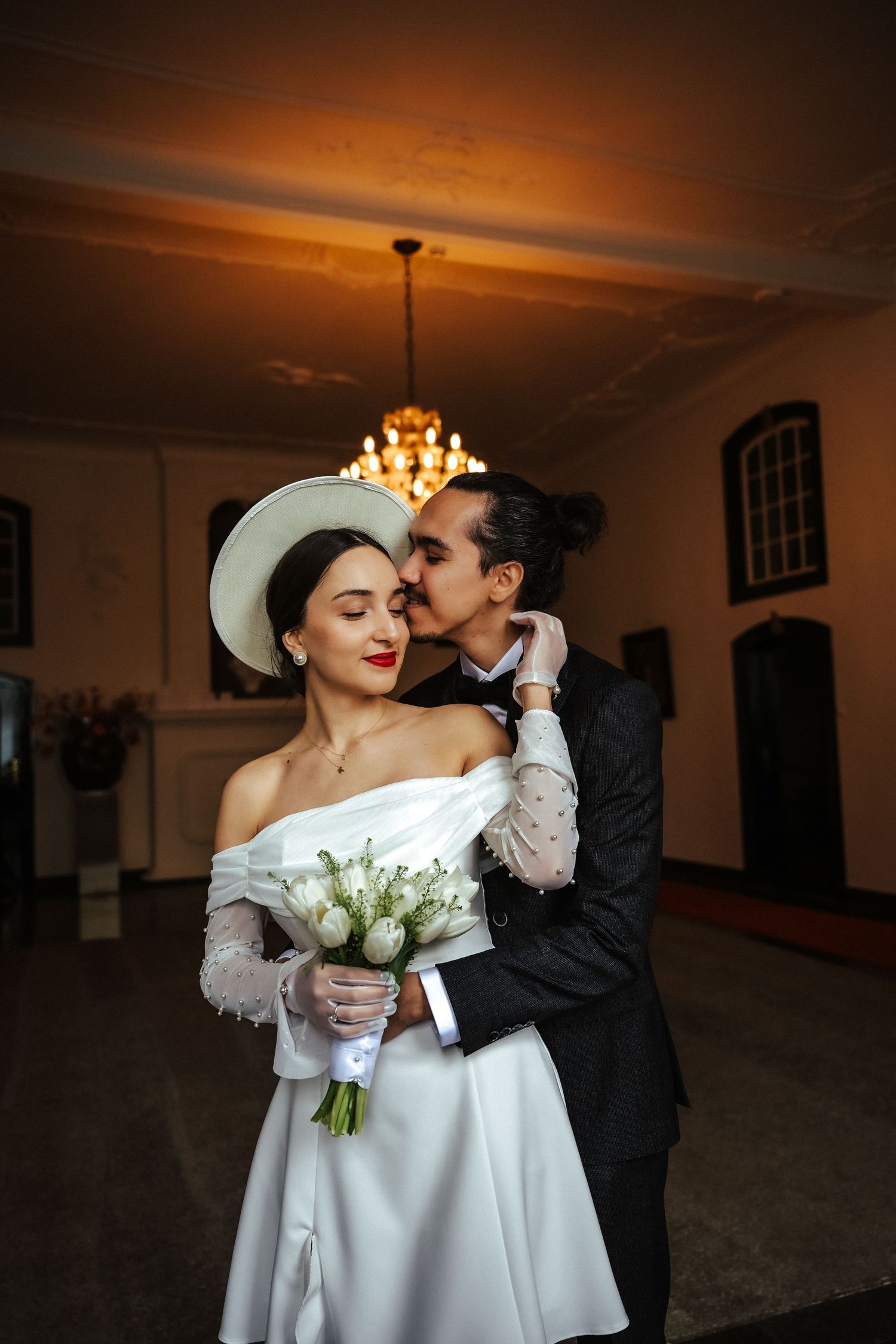 Romantic newlywed couple shares intimate moments in a dimly lit hallway with chandelier and white bridal bouquet.