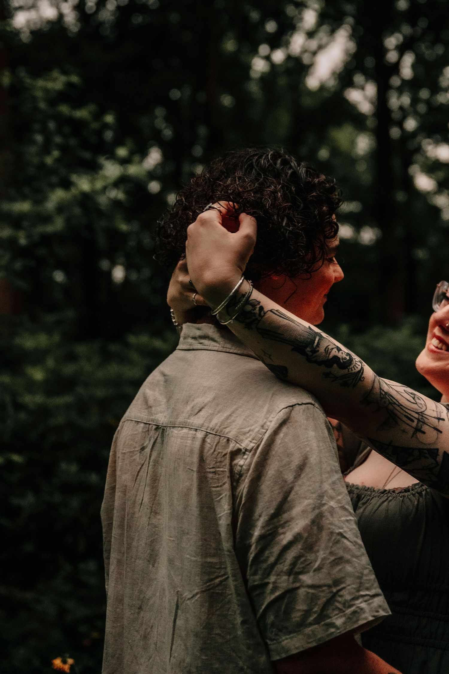 A couple embraces intimately outdoors in a moody forest setting with dark bokeh background.