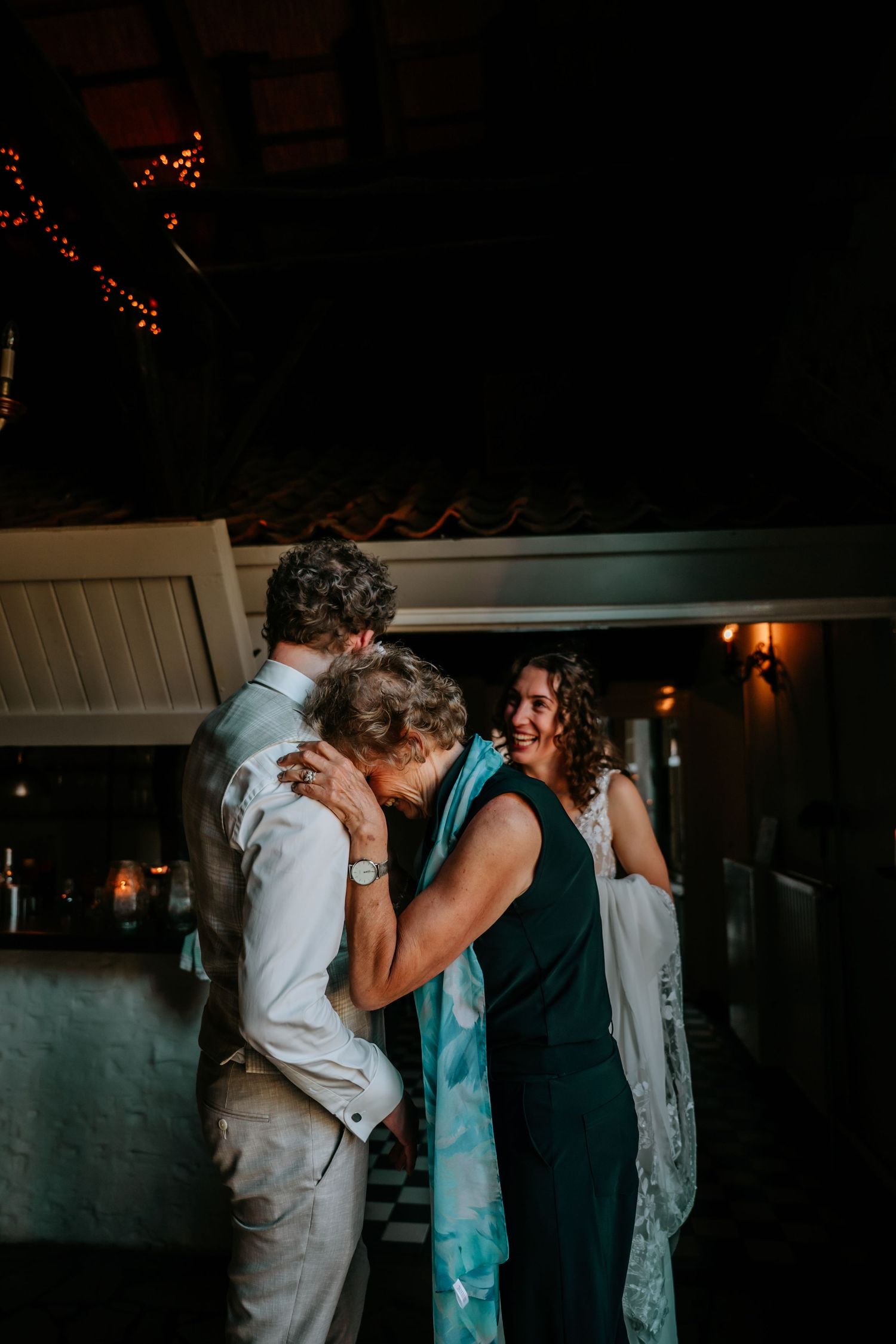 Wedding guests share an emotional embrace during an evening reception in a dimly lit venue.