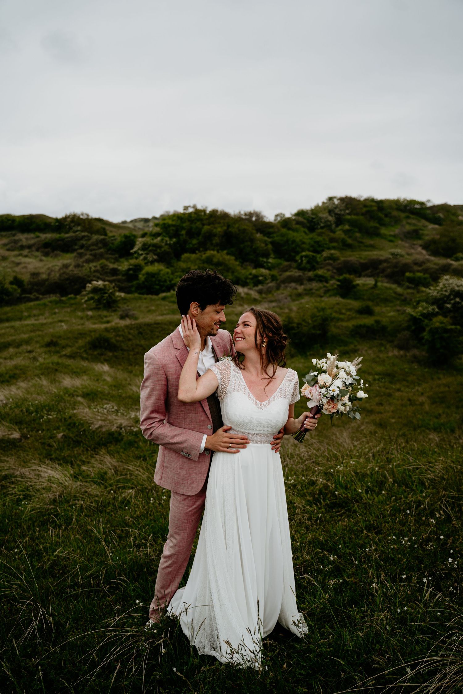 A couple in wedding attire share an intimate moment on a green hillside with the bride holding a white floral bouquet.
