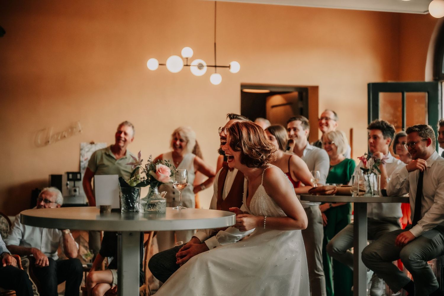 Wedding reception scene with guests seated at round tables under warm lighting.